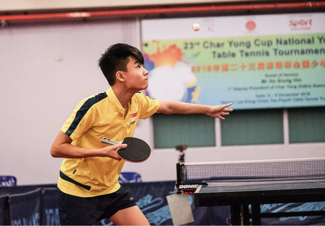 Young boy playing table tennis indoors, wearing a yellow sports shirt, preparing to hit the ball with his paddle.  He is called Nicholas and represented Singapore for overseas competitions.