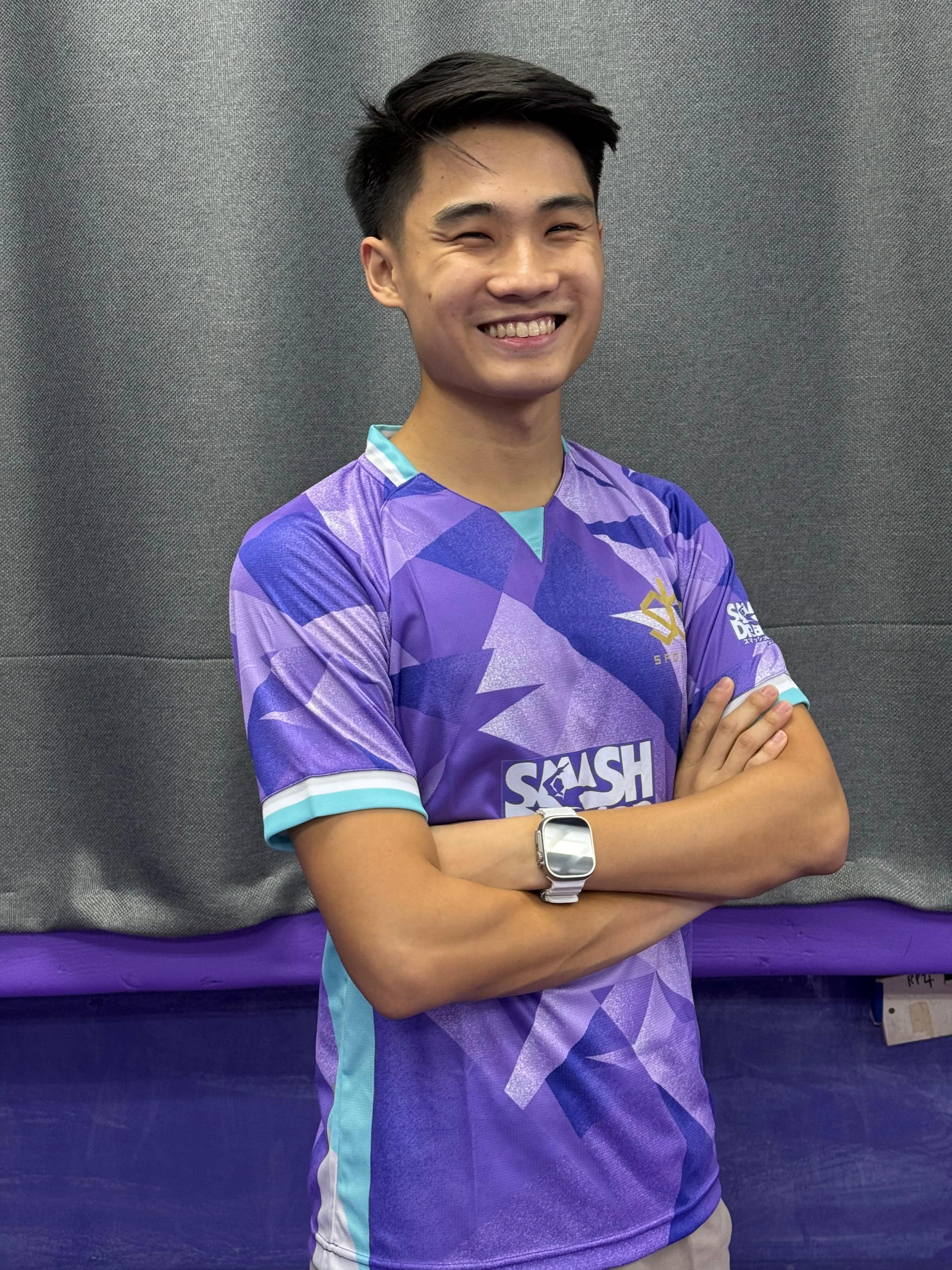 A young man with dark hair smiling, wearing a colorful sports jersey with purple, blue, and teal accents, crossing his arms, standing against a gray background.  He is called Nicholas and represented Singapore for overseas competitions.