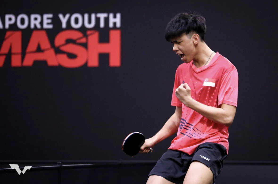 Young male table tennis player celebrating with a clenched fist, holding a paddle, wearing a pink shirt and black shorts, during a match at the Singapore Youth Smash event.