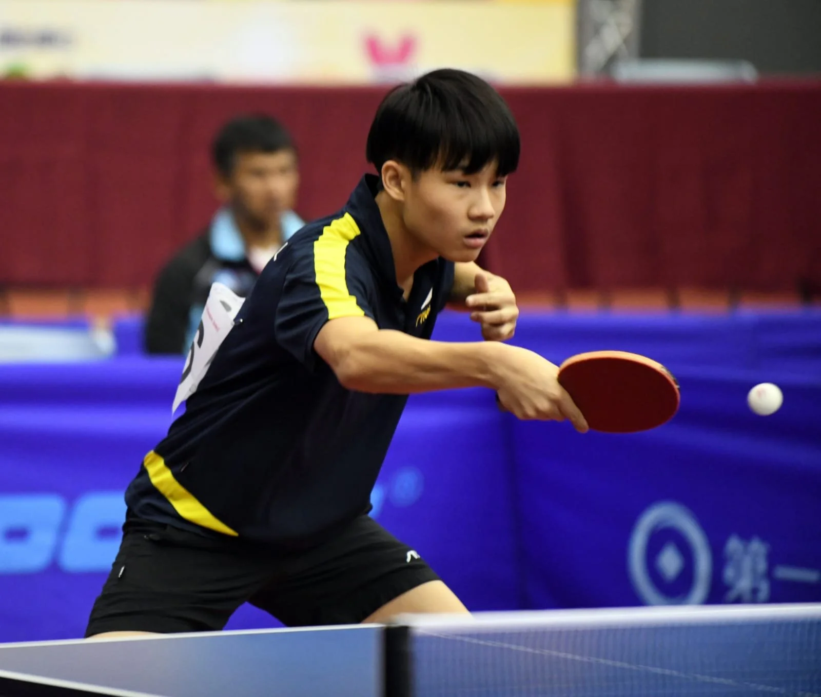 A young Asian boy playing table tennis, wearing a black sports uniform with yellow accents, focused on hitting the ball with a red paddle.  He is called Nicholas and represented Singapore for overseas competitions.