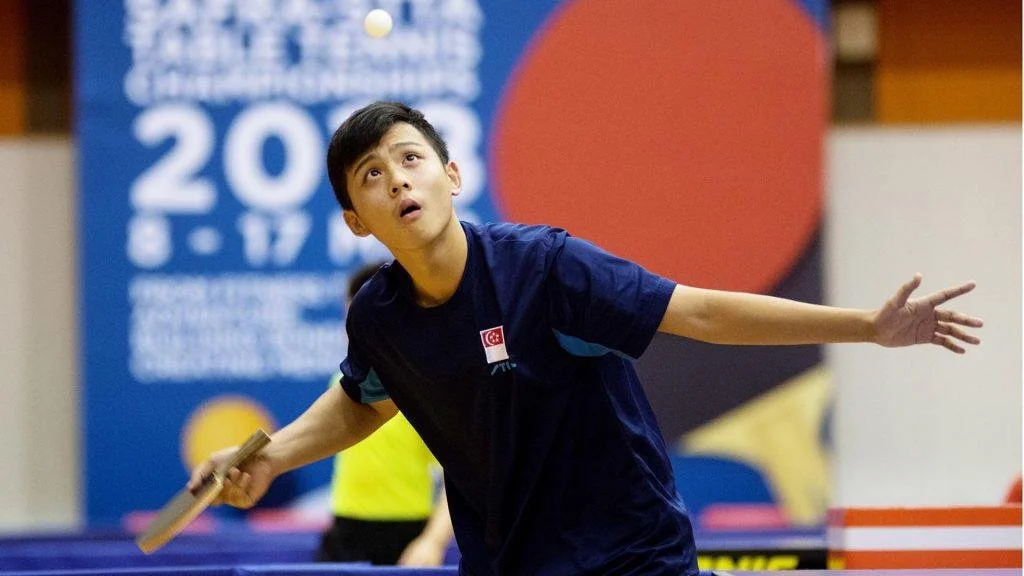 A young boy in a navy blue shirt playing table tennis, holding a paddle, with a focused expression, in an indoor sports facility.  He is called Andy and represented Singapore for overseas competitions.