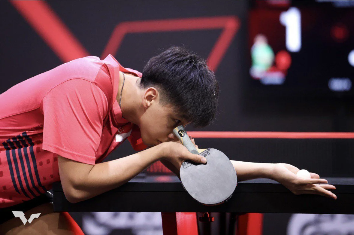 A male table tennis player in a pink shirt leans over a table, aiming a paddle and ball with his eye close to the paddle, preparing to serve or make a shot.  He is called Jayden and represented Singapore for overseas competitions.