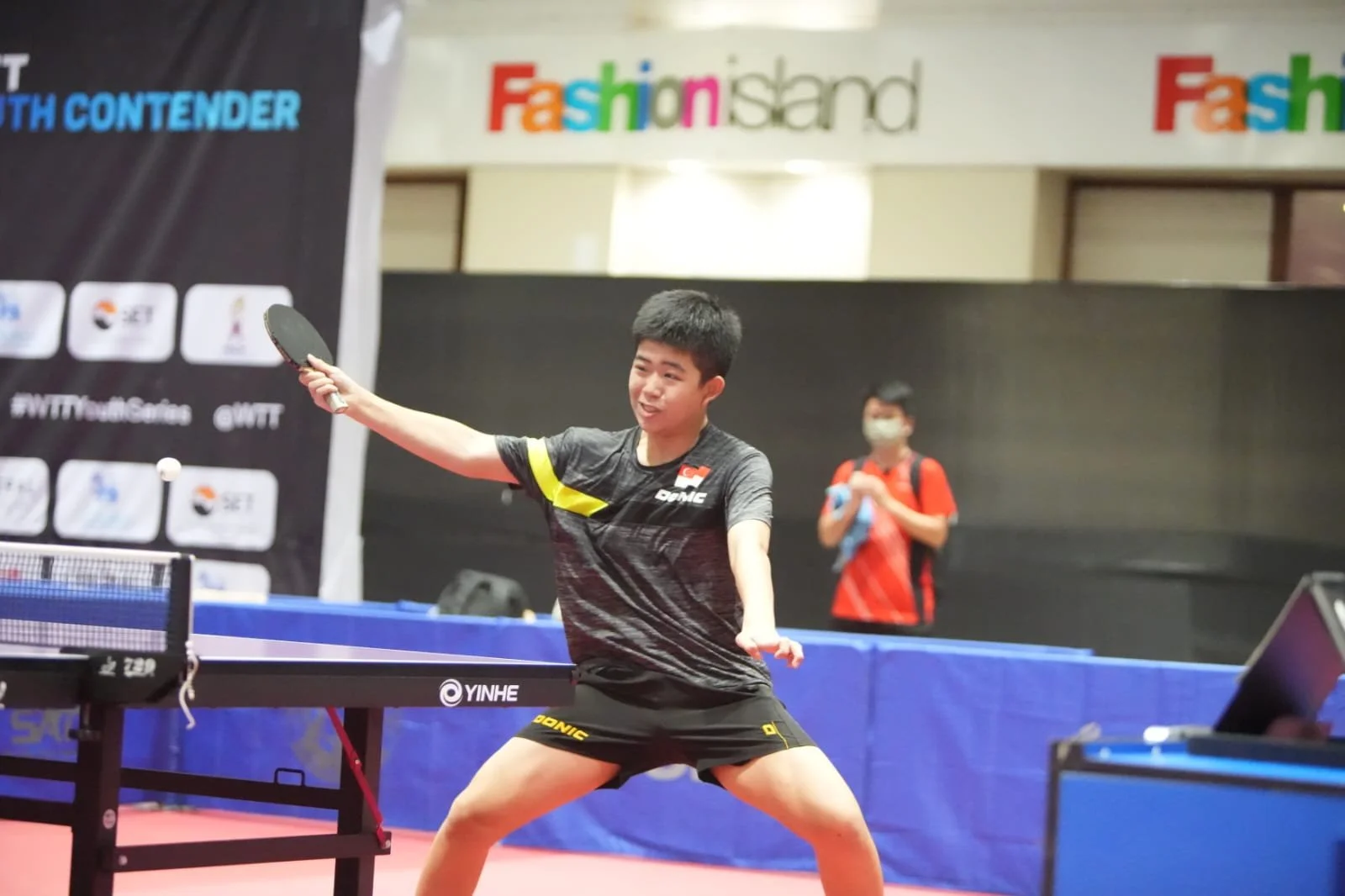 A young male table tennis player in a black and yellow uniform is in a ready stance, preparing to hit the ball during a match at a table tennis facility.  He is called Aron and represented Singapore for overseas competitions.