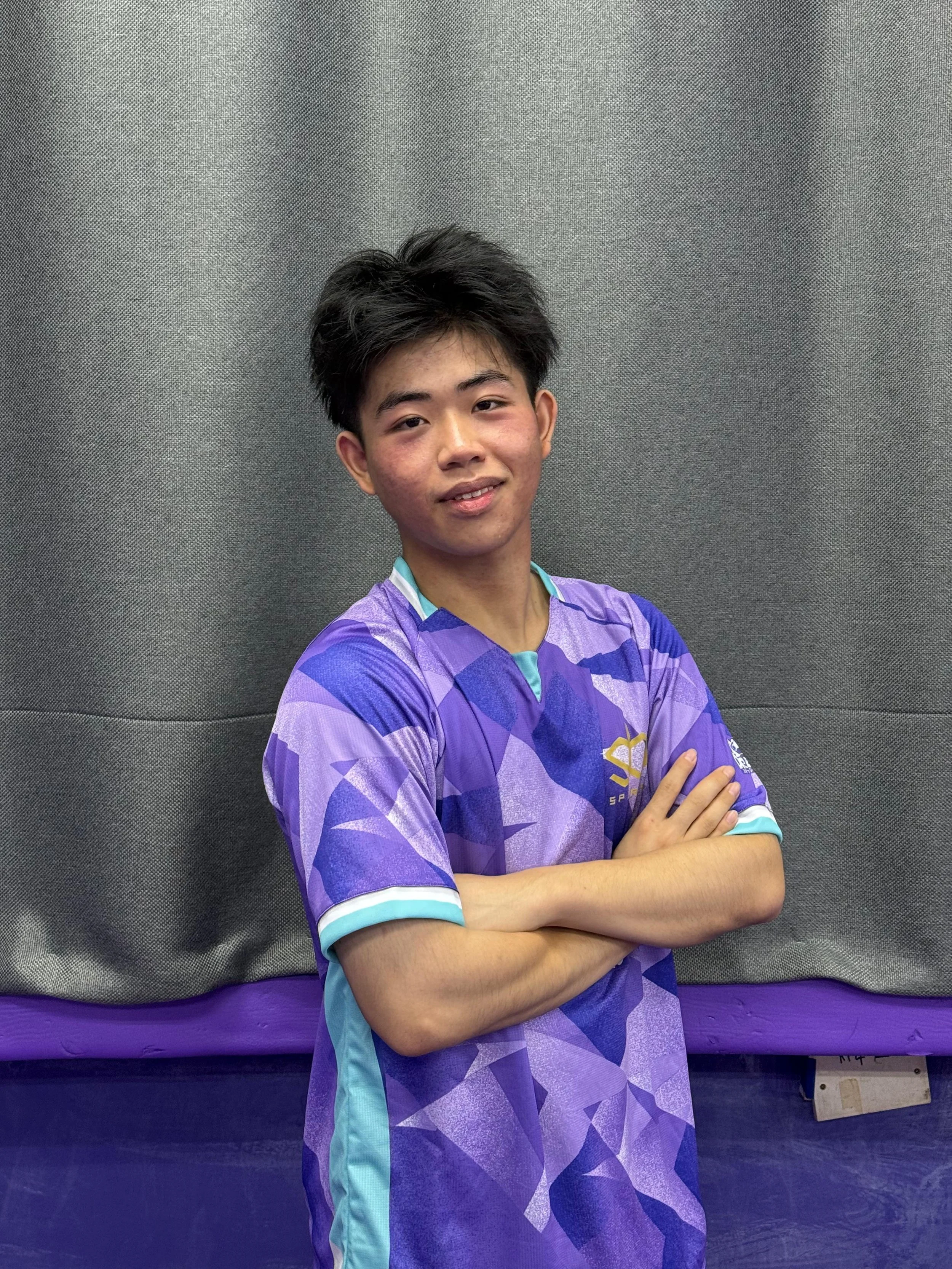 A young man with dark, tousled hair and a slight smile, standing with arms crossed against a gray background, wearing a purple and blue patterned sports jersey. He is called Aron and represented Singapore for overseas competitions.