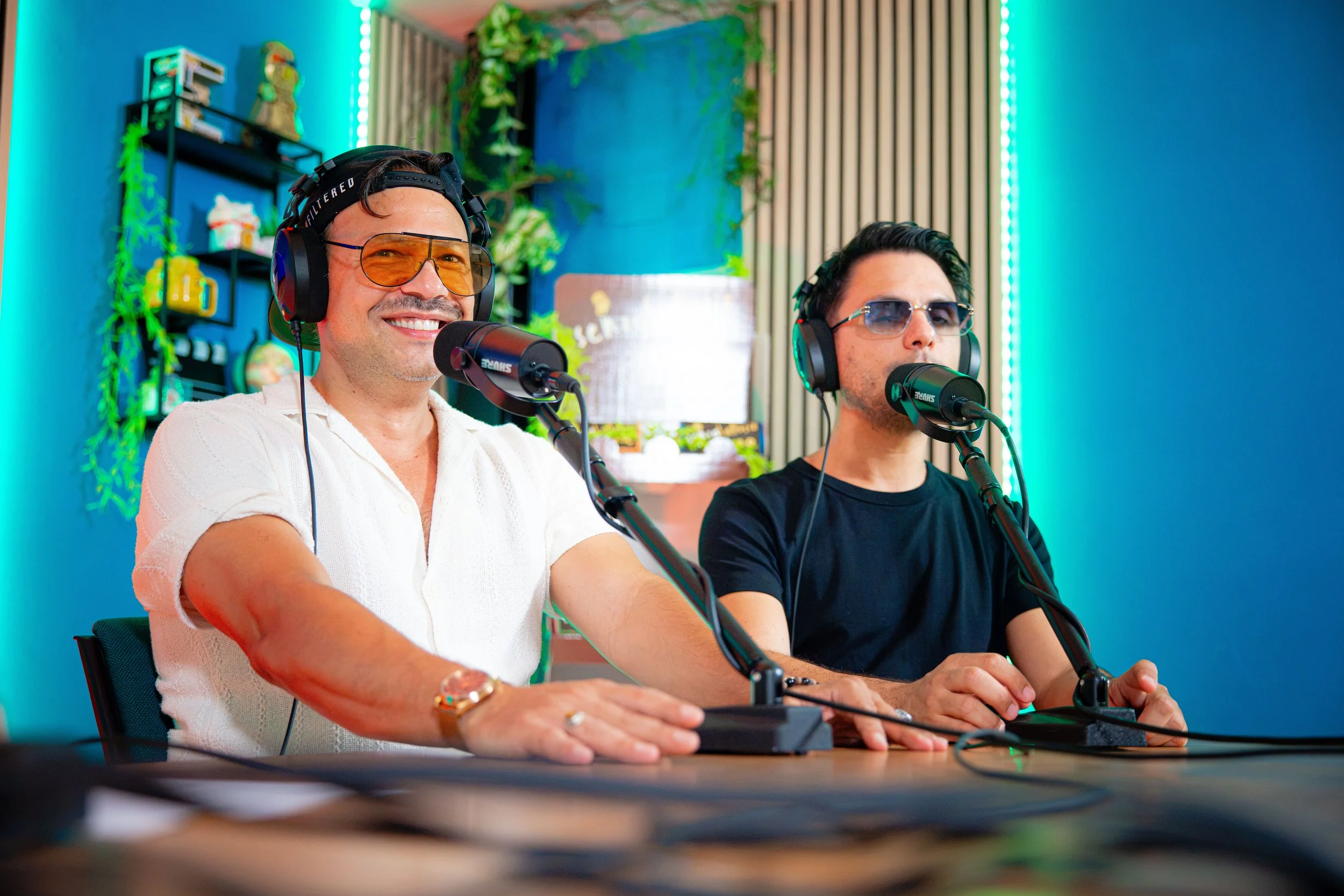 Due uomini seduti a un tavolo durante una registrazione radiofonica, indossano cuffie e parlano davanti a microfoni in uno studio decorato con piante e luci blu.