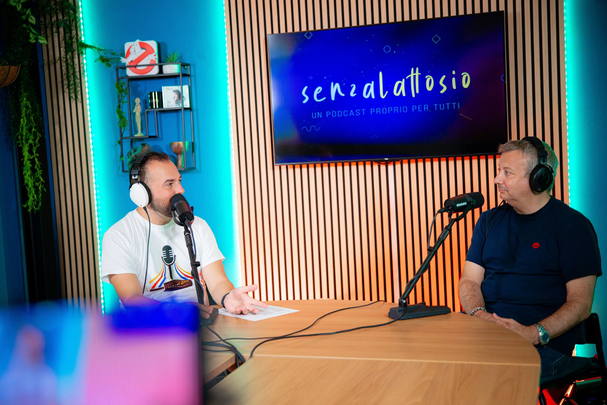 Due uomini seduti a un tavolo durante una conversazione, indossano cuffie e parlano in un podcast, con uno sfondo di luci soffuse e una grande schermata con la scritta 'senza lattosio' e 'UN PODCAST PROPRIO PER TUTTI'.