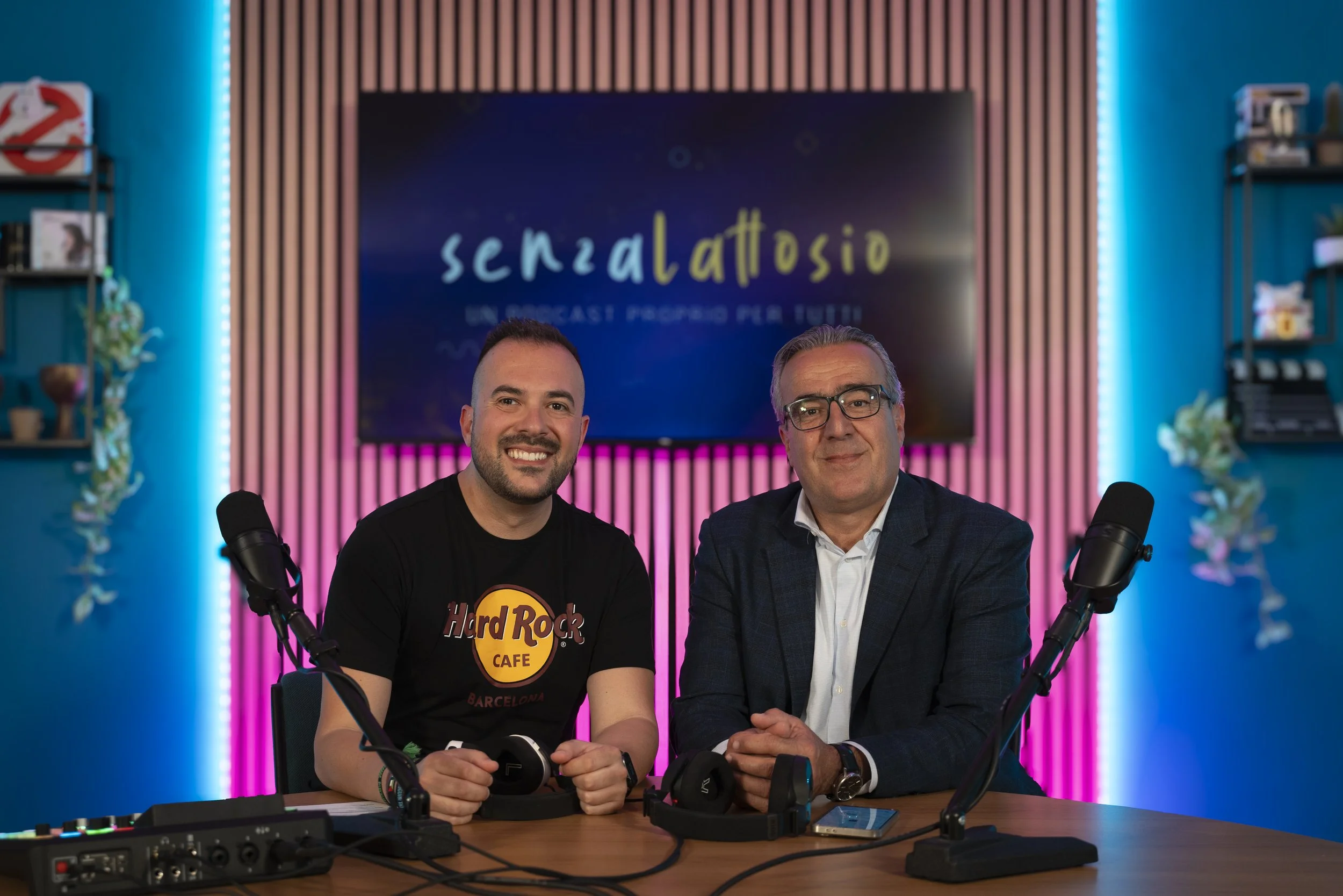 Due uomini sono seduti a una scrivania davanti a microfoni di registrazione, in uno studio con pareti decorate con luci LED blu e rosa. Sullo sfondo si vede un grande schermo con il testo 'Senza Attioso'.