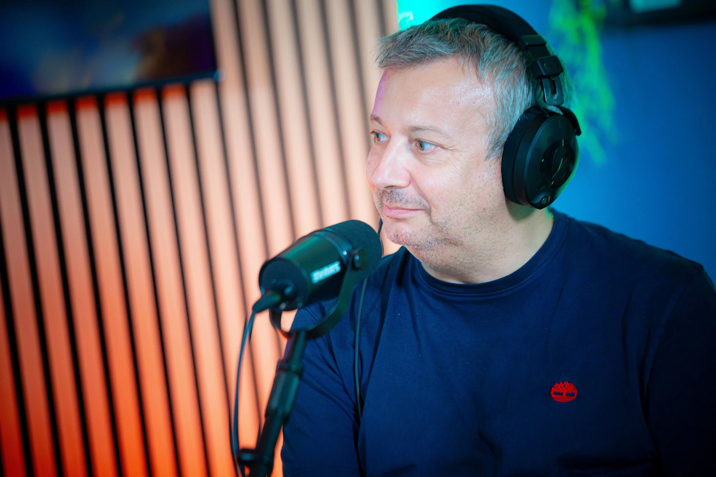 Uomo con capelli grigi e occhi azzurri che indossa cuffie nere e parla davanti a un microfono in uno studio di registrazione, sfondo con pannelli di legno e luci colorate.