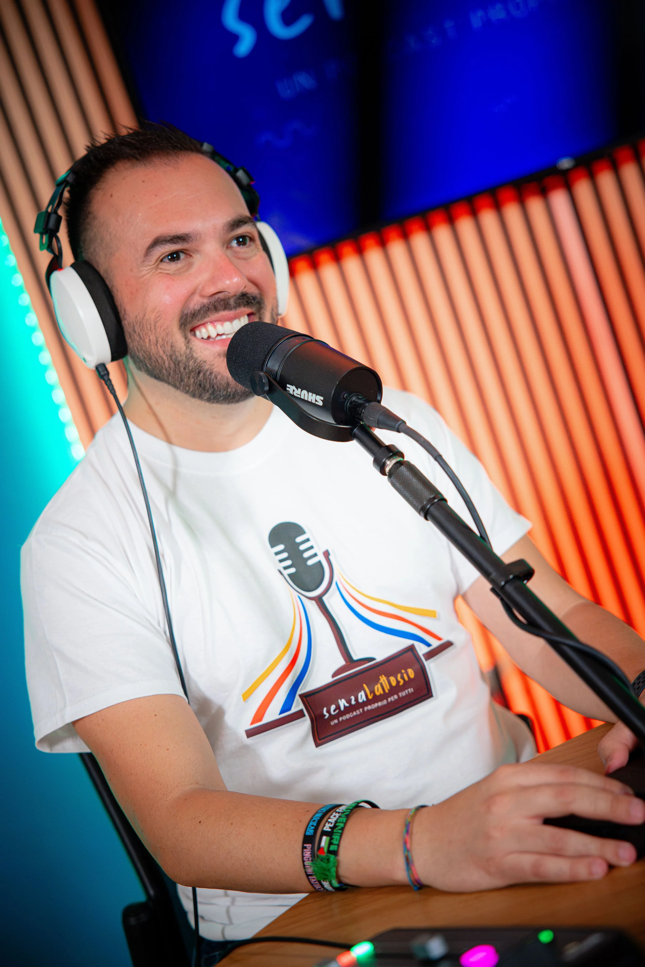 Un uomo sorridente con barba e capelli corti, indossa cuffie bianche e neri e una maglietta bianca con un logo colorato, mentre parla davanti a un microfono in uno studio di registrazione. Sullo sfondo ci sono pannelli di legno e uno schermo blu.