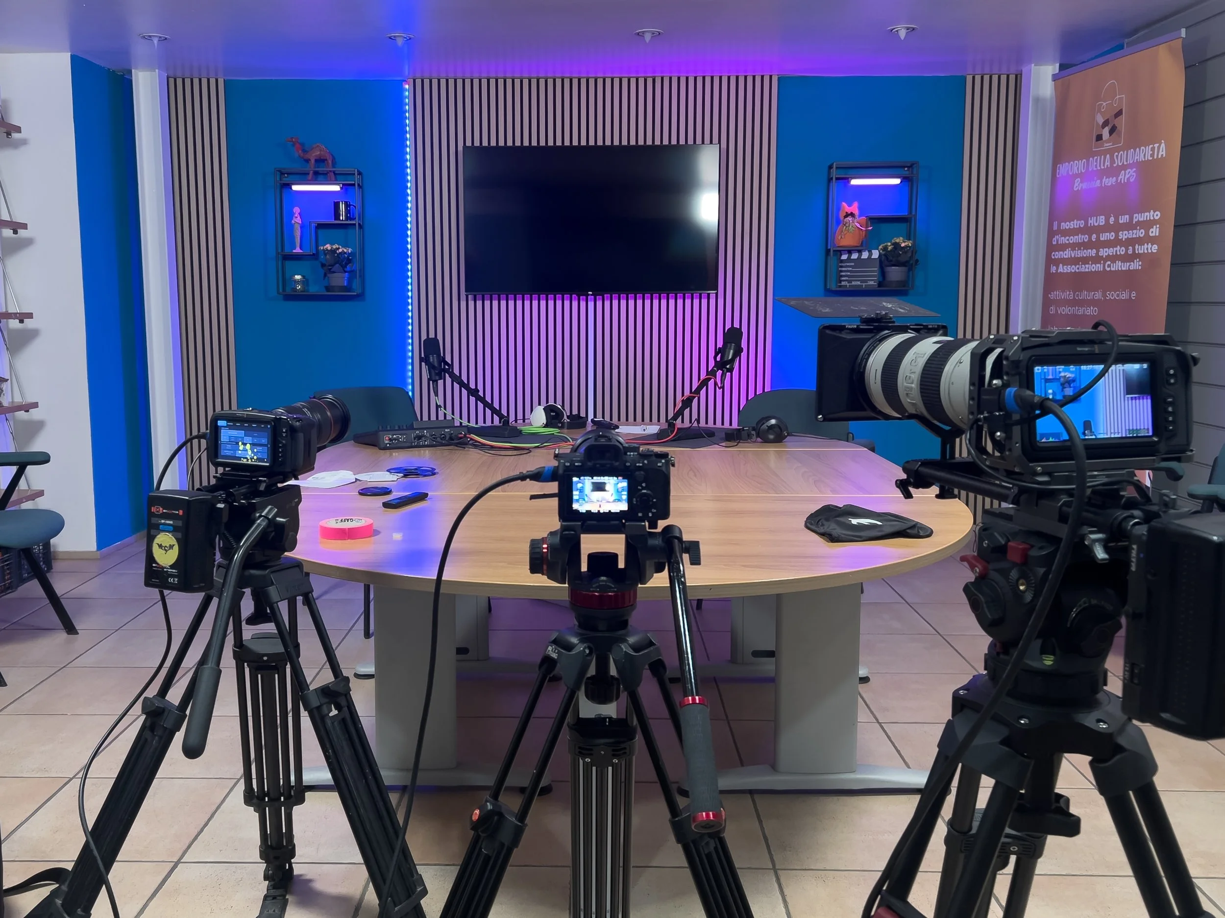 Sala di riunioni con telecamere e attrezzature per registrazioni, grande tavolo rotondo, schermo TV sulla parete, luci colorate, scaffali con oggetti decorativi e banner di informazione in italiano.