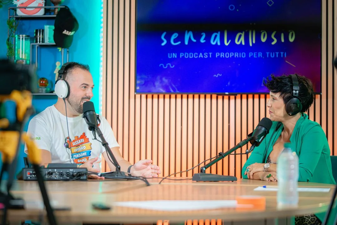 Due persone parlano in un podcast, sedute a un tavolo con microfoni, casco audio e apparecchiature di registrazione, in uno studio con arredi in legno e una grande schermata sullo sfondo che dice "senzatattosio" e "un podcast proprio per tutti".