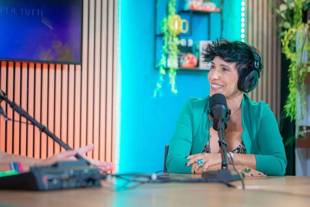 Desirè Manca sorridente con cuffie ascolta parlando in un podcast o in una trasmissione radio in uno studio colorato con pareti di legno e piante.