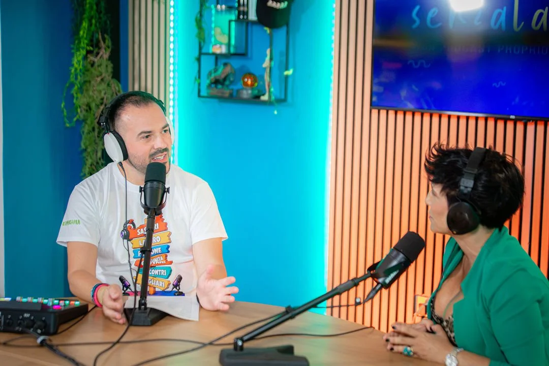 Due persone sedute a un tavolo in una registrazione radiofonica, parlando in microfoni e indossando cuffie, in un ambiente ben illuminato con pareti in legno e una parete blu.