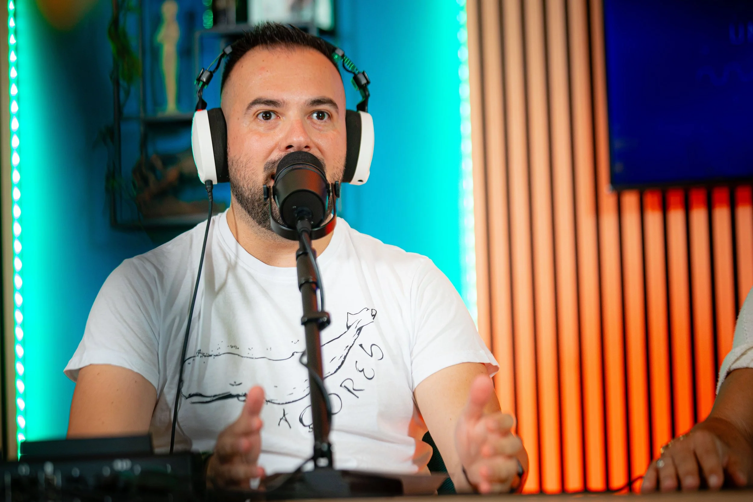 Uomo con capelli corti e barba, che indossa cuffie grandi e parla davanti a un microfono in uno studio di registrazione, con schermi e pareti colorate sullo sfondo.
