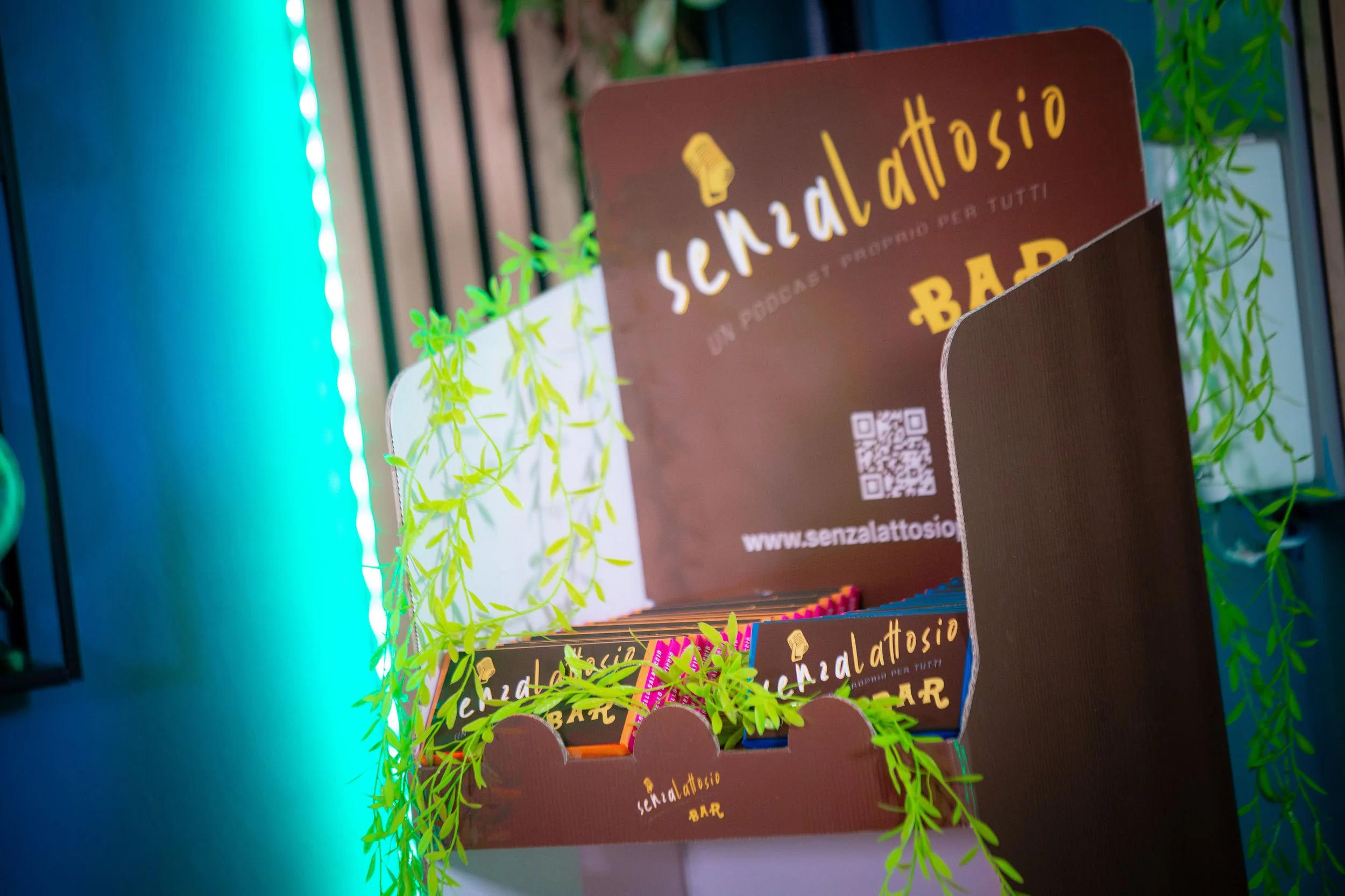 Un cartellone pubblicitario per un podcast 'Senzalattosio' con un'illustrazione di un microfono e decorazioni di foglie verdi, sullo sfondo luci blu e ambientazione moderna.