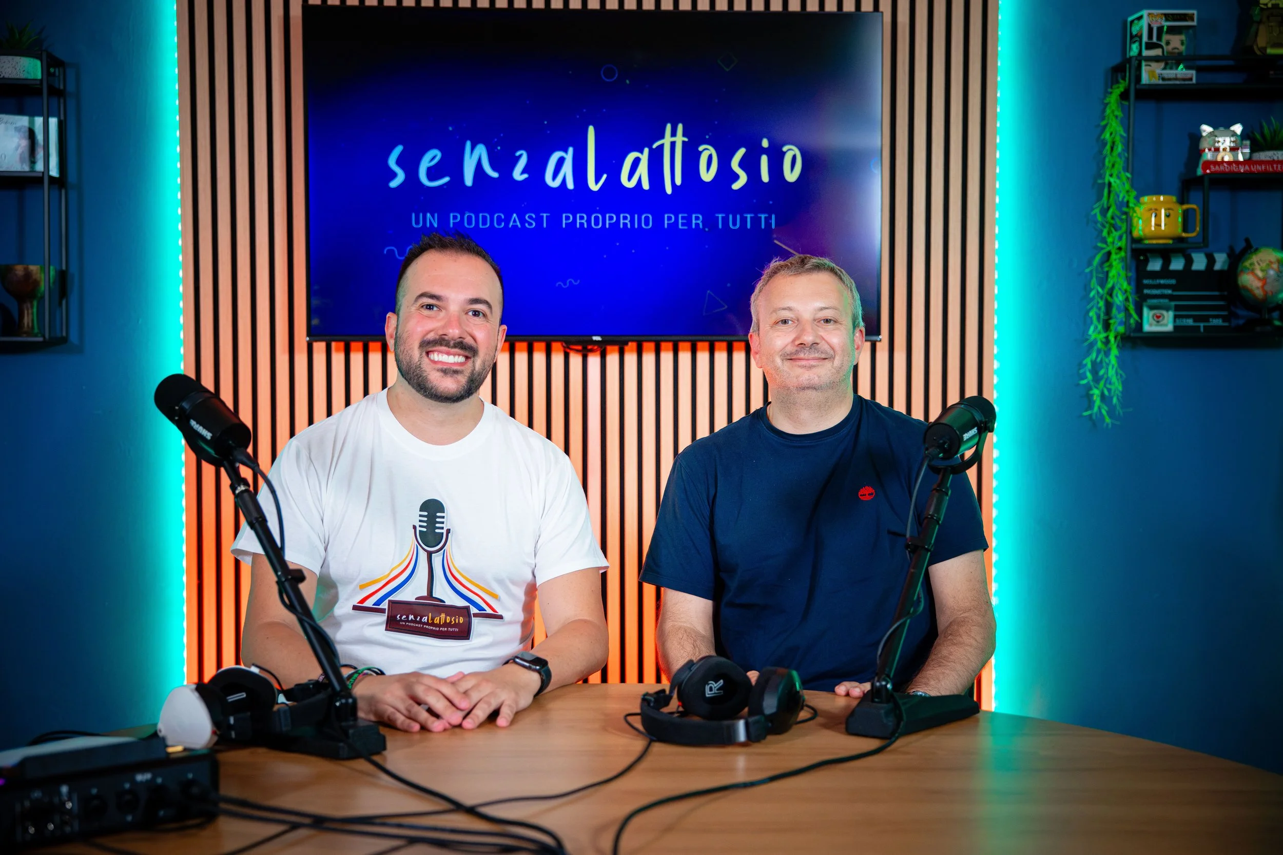Due uomini sorridenti seduti a un tavolo davanti a microfoni, in uno studio di registrazione o podcast, con uno sfondo digitale che mostra il titolo 'Senza Lattosio' e la scritta 'Un podcast proprio per tutti', con scaffali con oggetti decorativi.