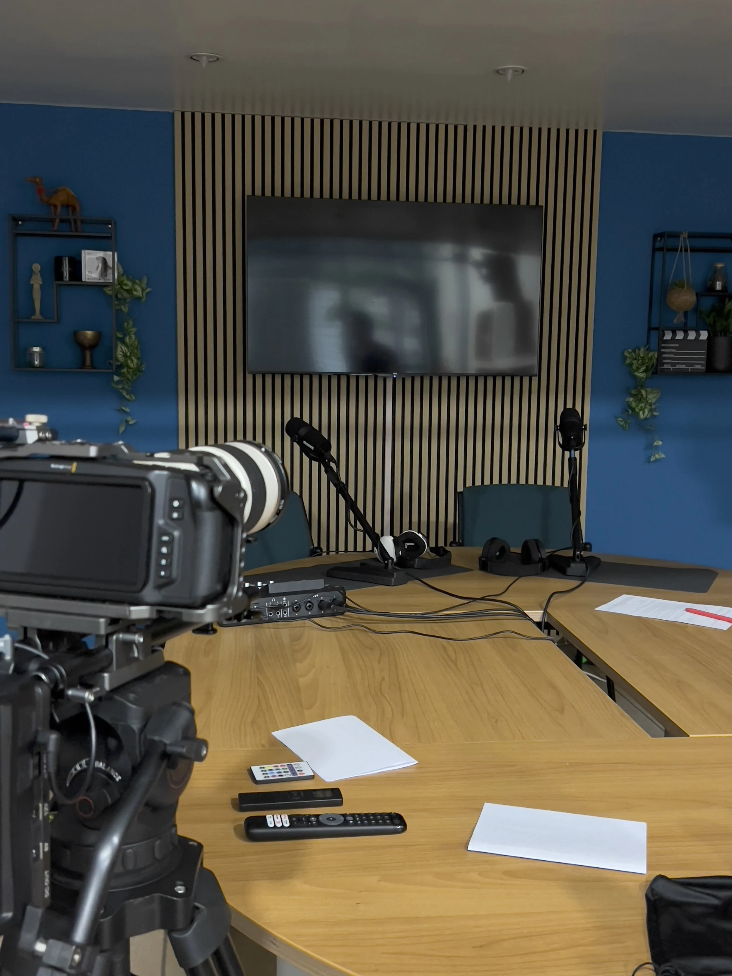 Sala riunioni con telecamera, microfoni e schermo grande, con pareti blu e scaffali decorativi.