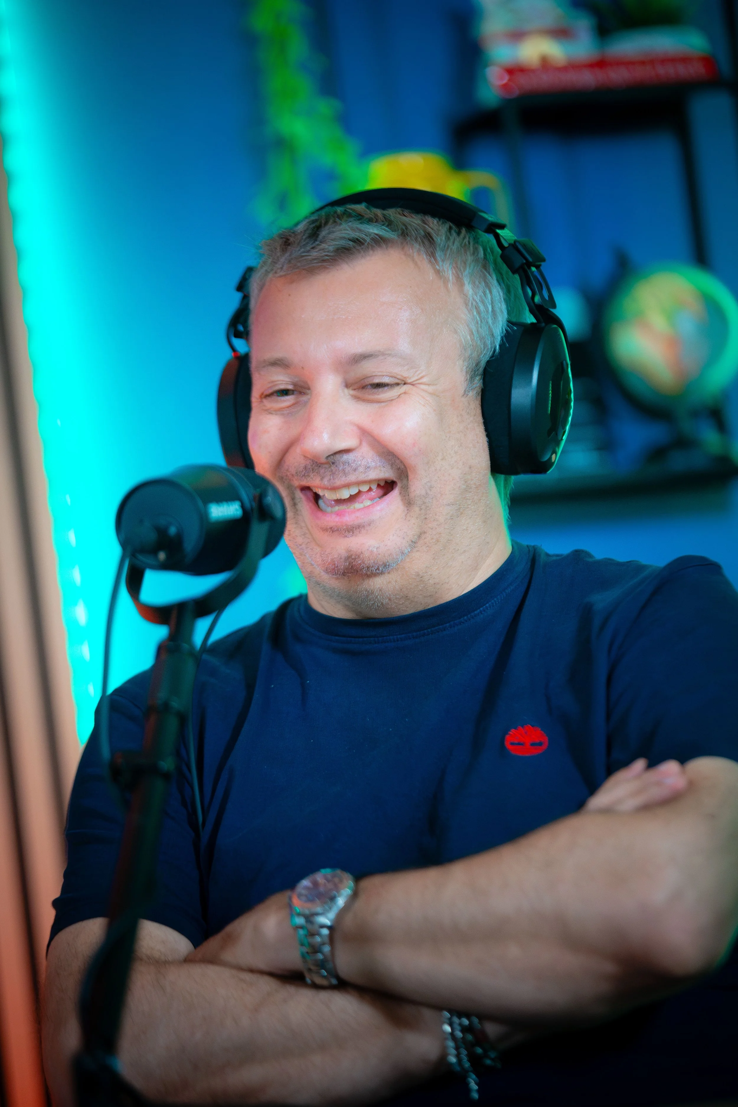 Uomo che indossa cuffie parlando in microfono in uno studio, sfondo con oggetti decorativi e colori vivaci.