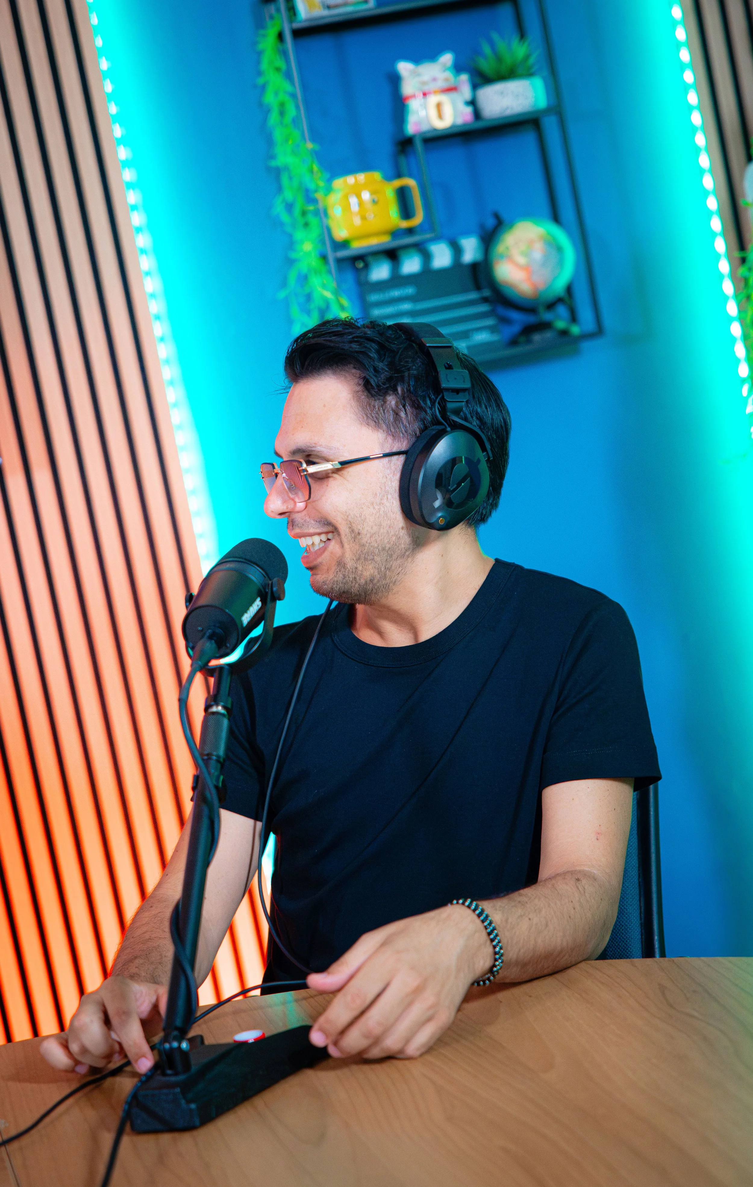 Uomo sorridente con occhiali e cuffie, seduto a un tavolo, che parla in un microfono in uno studio colorato con scaffali decorativi sullo sfondo.