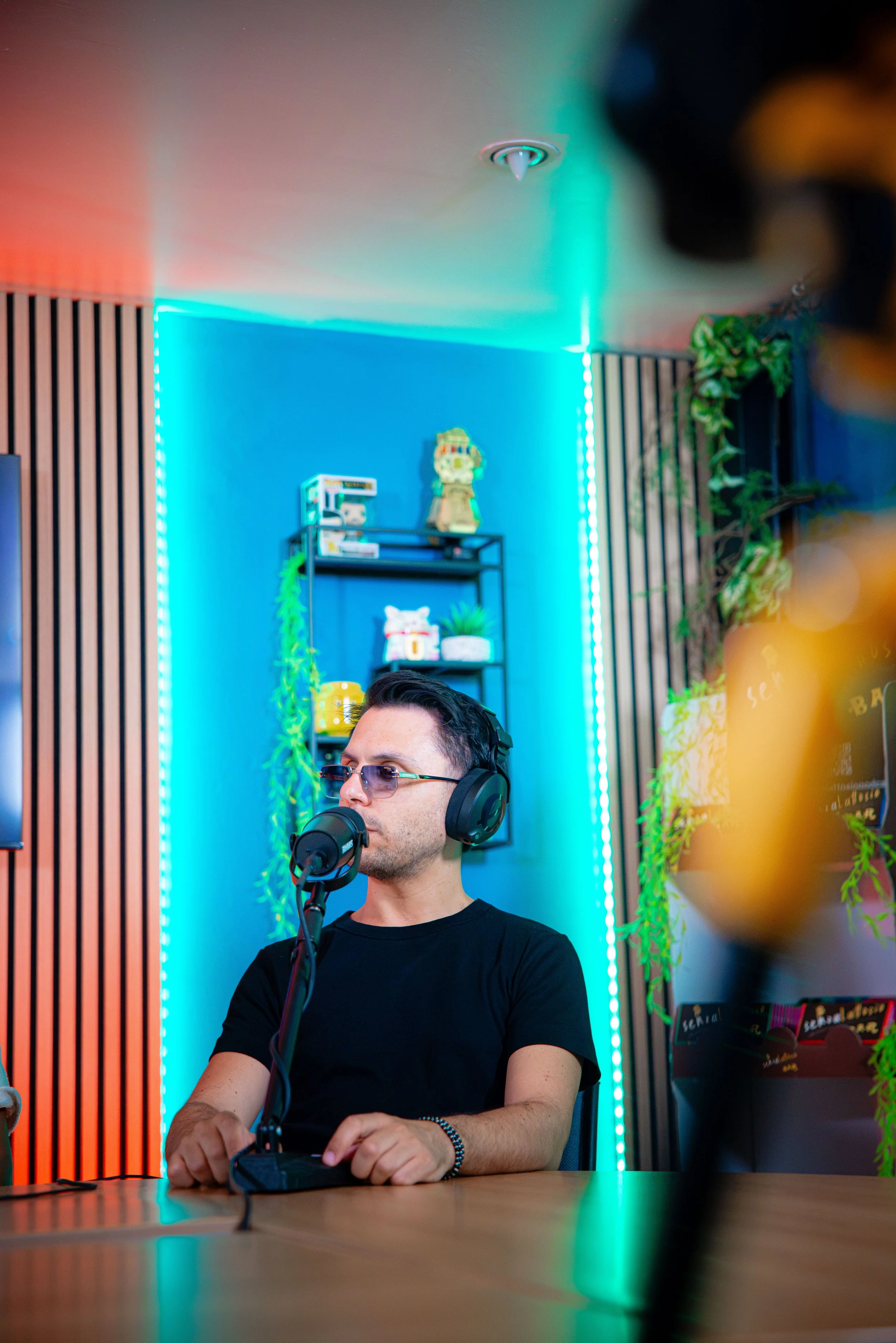 Un uomo con occhiali e cuffie sta parlandando in uno studio di registrazione o in un podcast, con sfondo di luci blu e verde, scaffali con oggetti decorativi e piante vere.