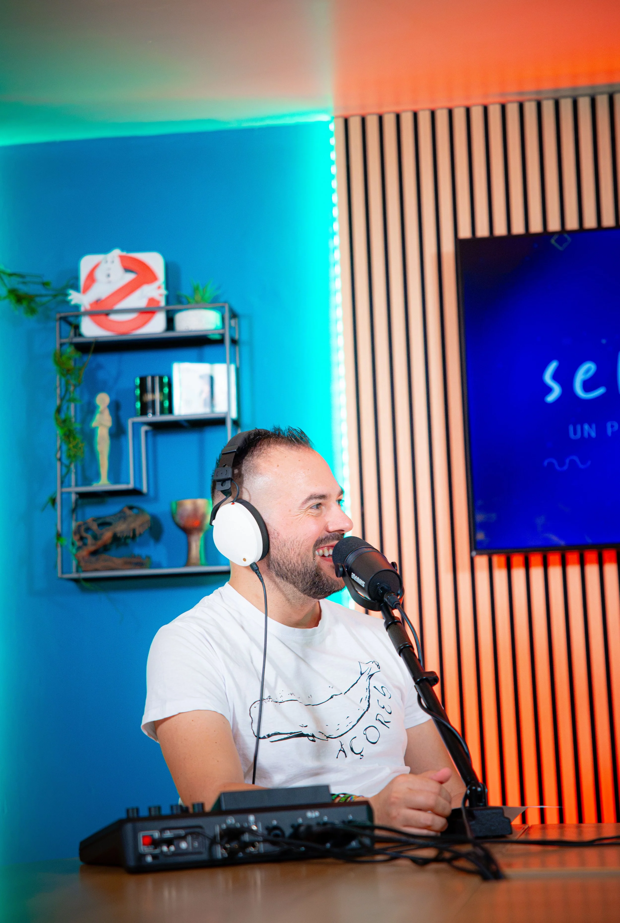 Un uomo con cuffie bianche e microfono ascolta e sorride, seduto davanti a un mixer audio, all'interno di uno studio con pareti colorate e decorazioni.