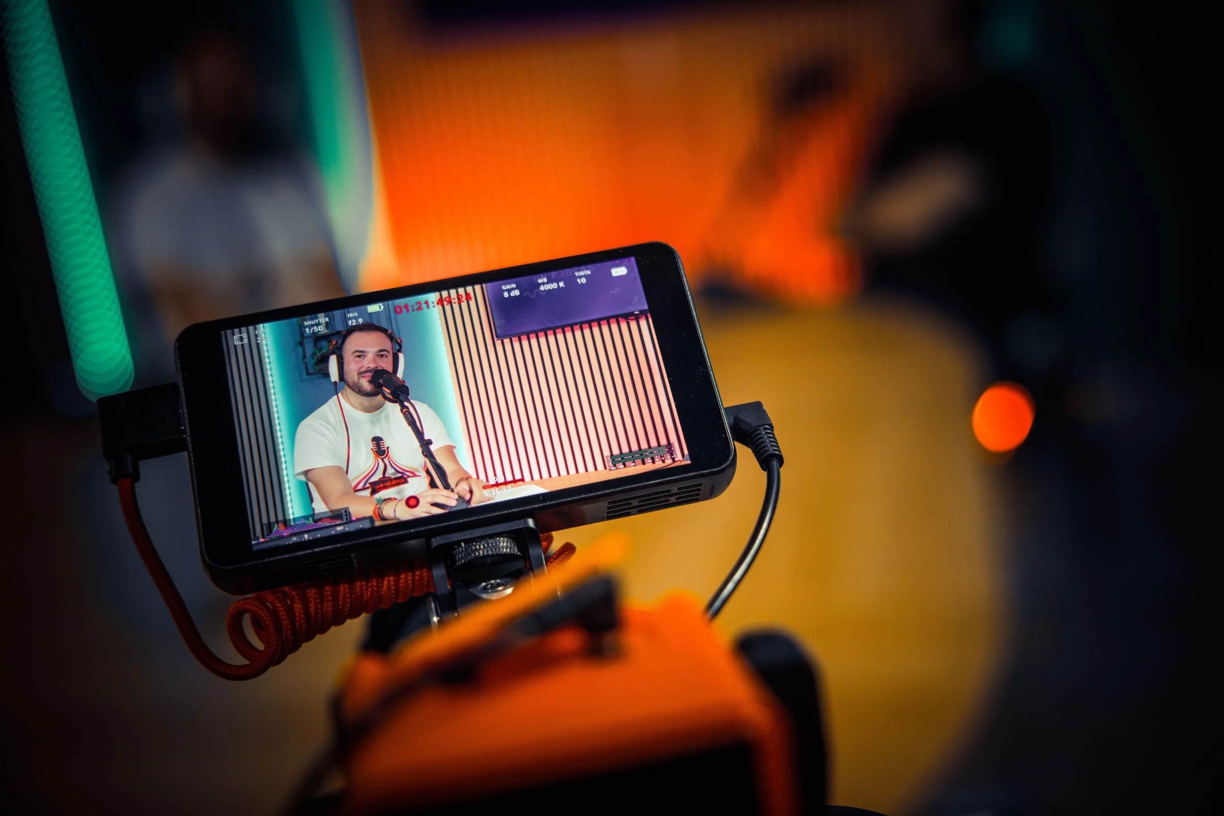 Uno schermo di telecamera mostra un uomo con cuffie che parla al microfono durante una registrazione in uno studio con luci colorate.