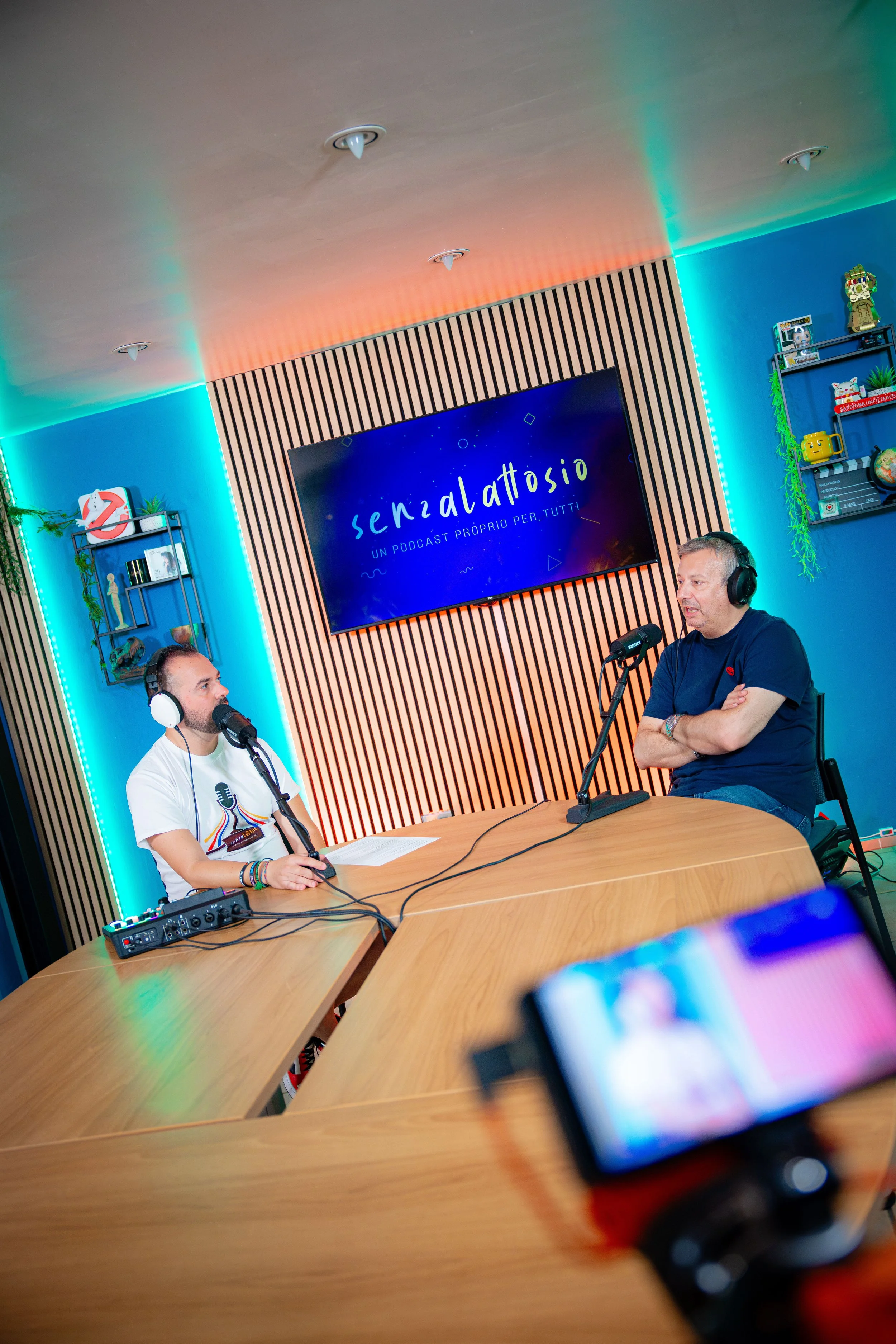 Due uomini seduti a un tavolo in una stanza che sembra un podcast studio, con uno schermo dietro di loro che mostra il testo 'senza lattosio' e 'un podcast proprio per tutti'. Uno indossa cuffie e parla al microfono, l'altro ascolta, mentre una video