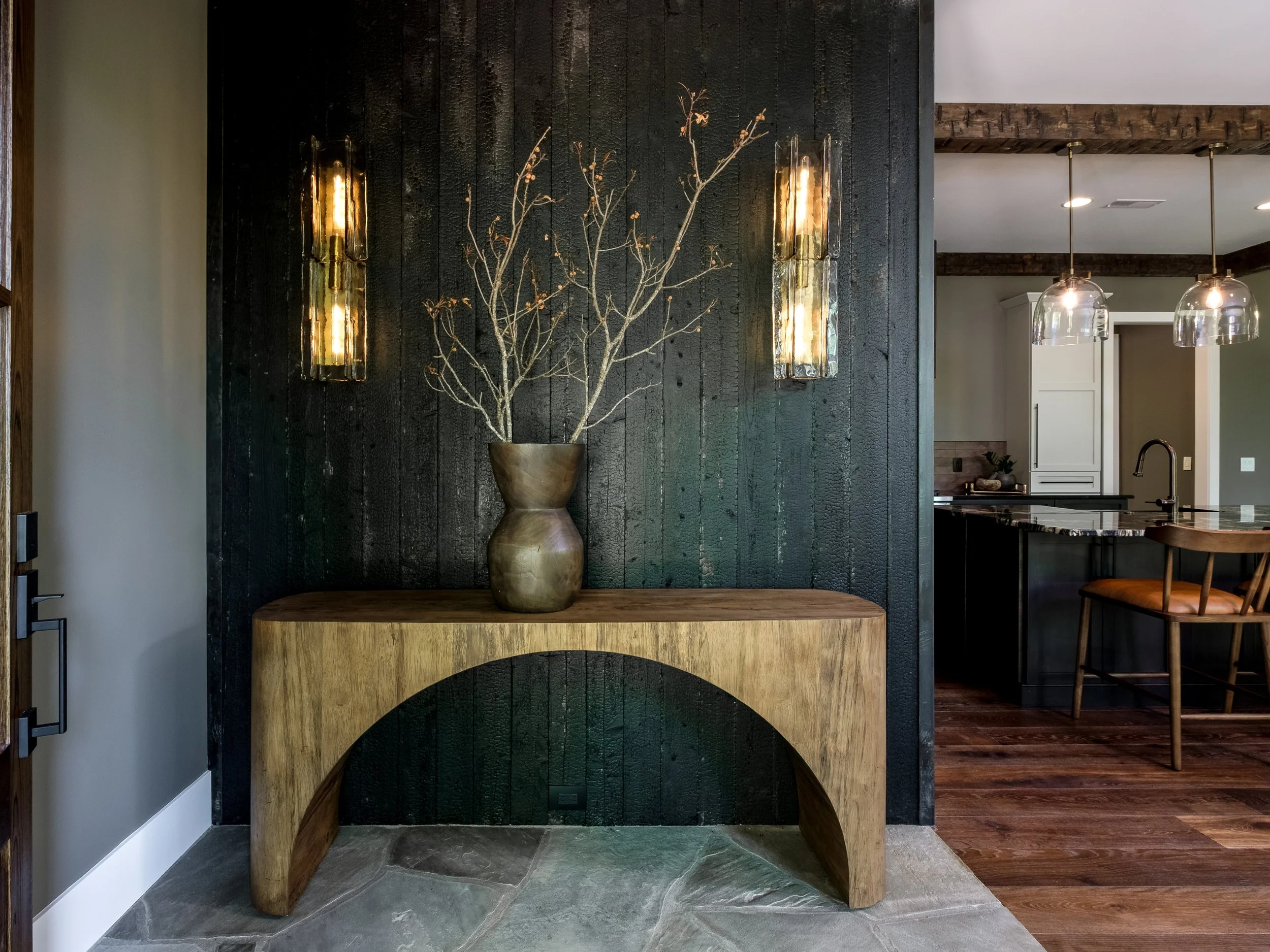 A wooden console table with a large bronze vase containing bare branches is positioned against a dark textured wall. Two modern wall sconces with warm lighting flank the vase. In the background, a kitchen with pendant lights over a dark island and wooden flooring is visible.