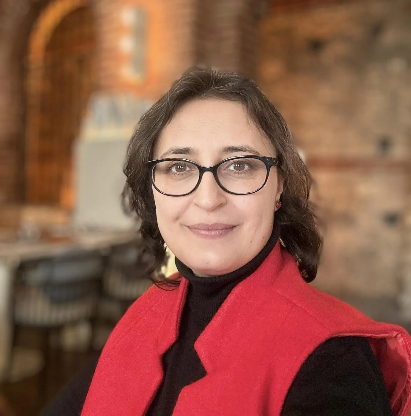A woman with short, wavy brown hair and glasses, smiling with a neutral expression. She is wearing a red vest over a black turtleneck sweater, and earrings. The background features a brick wall and blurred furniture.
