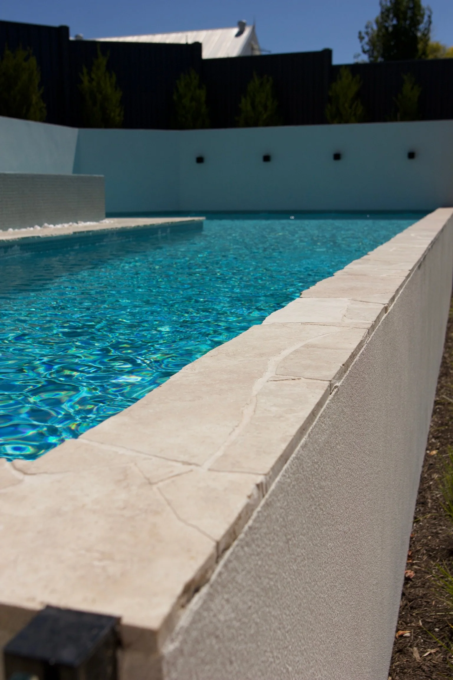 A modern outdoor swimming pool with clear blue water, surrounded by a white stone deck and a tall white wall, under a clear daytime sky.