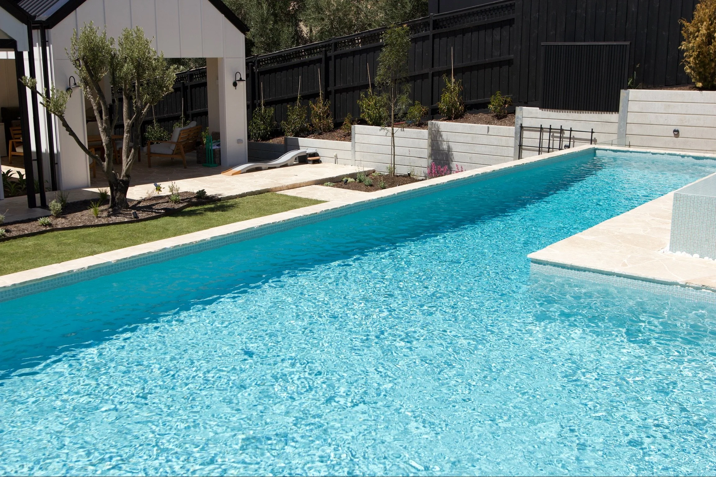 Backyard with a rectangular swimming pool, lounge chairs, trees, plants, and a house with a porch and outdoor seating.