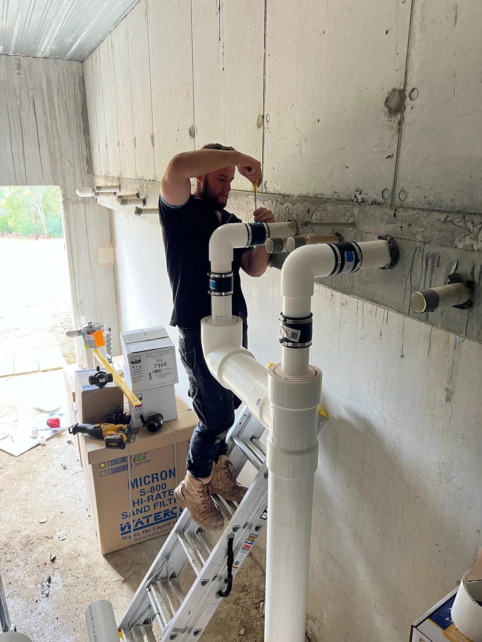 A man working on plumbing installation with PVC pipes inside a building under construction, standing on a ladder