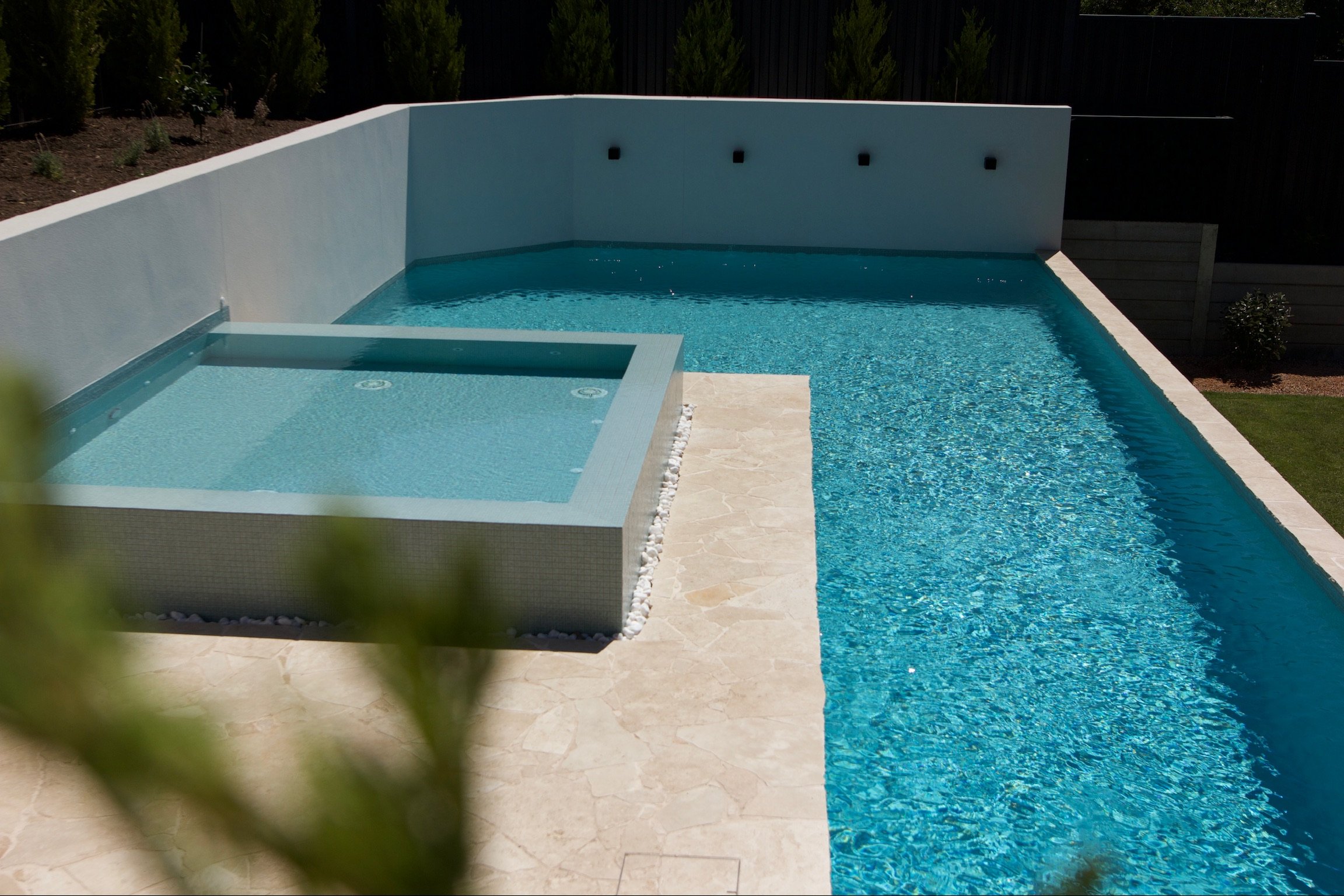 A backyard swimming pool with a small hot tub, surrounded by a stone deck and white wall, with a dark fence and greenery in the background.