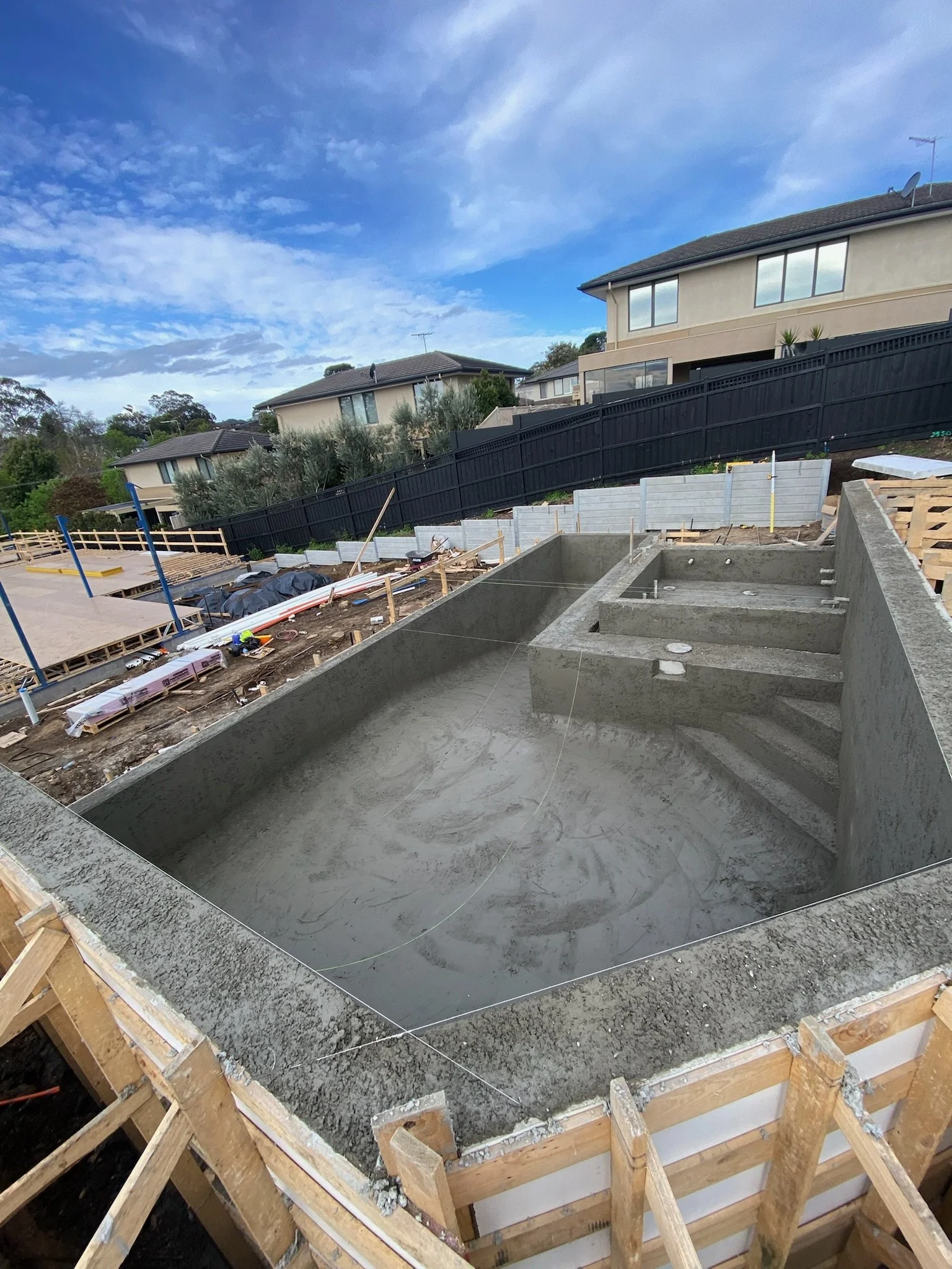 A swimming pool under construction in a backyard, with concrete and wooden forms, neighboring houses, and a partly cloudy sky.