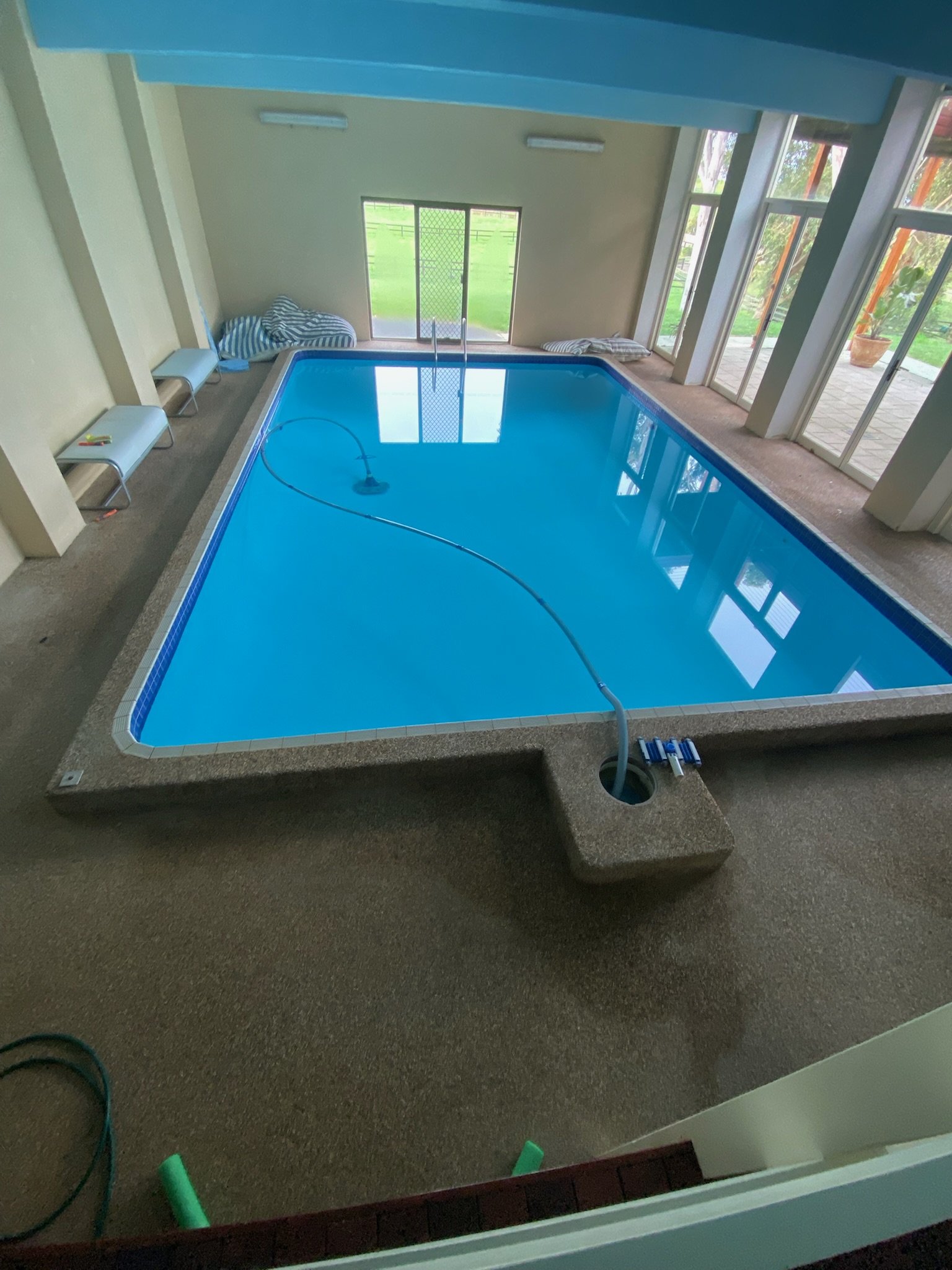 Indoor swimming pool with blue water, surrounded by beige walls and large windows. Pool cleaning equipment is visible, and sunlight streams in through the windows.