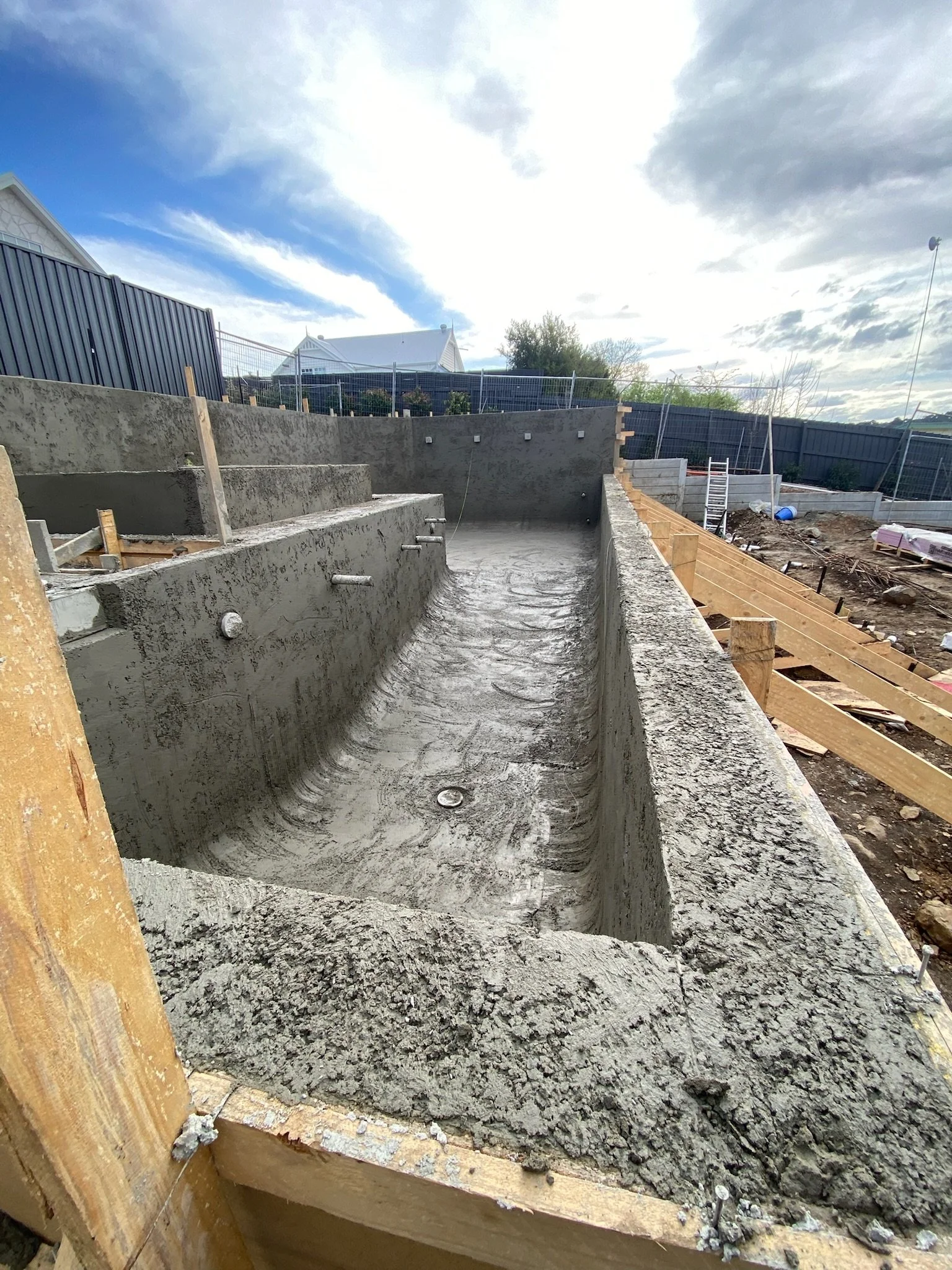 A concrete swimming pool in the process of construction, with the shell poured and prepared for finishing, surrounded by a construction site with dirt, wooden forms, and building materials.