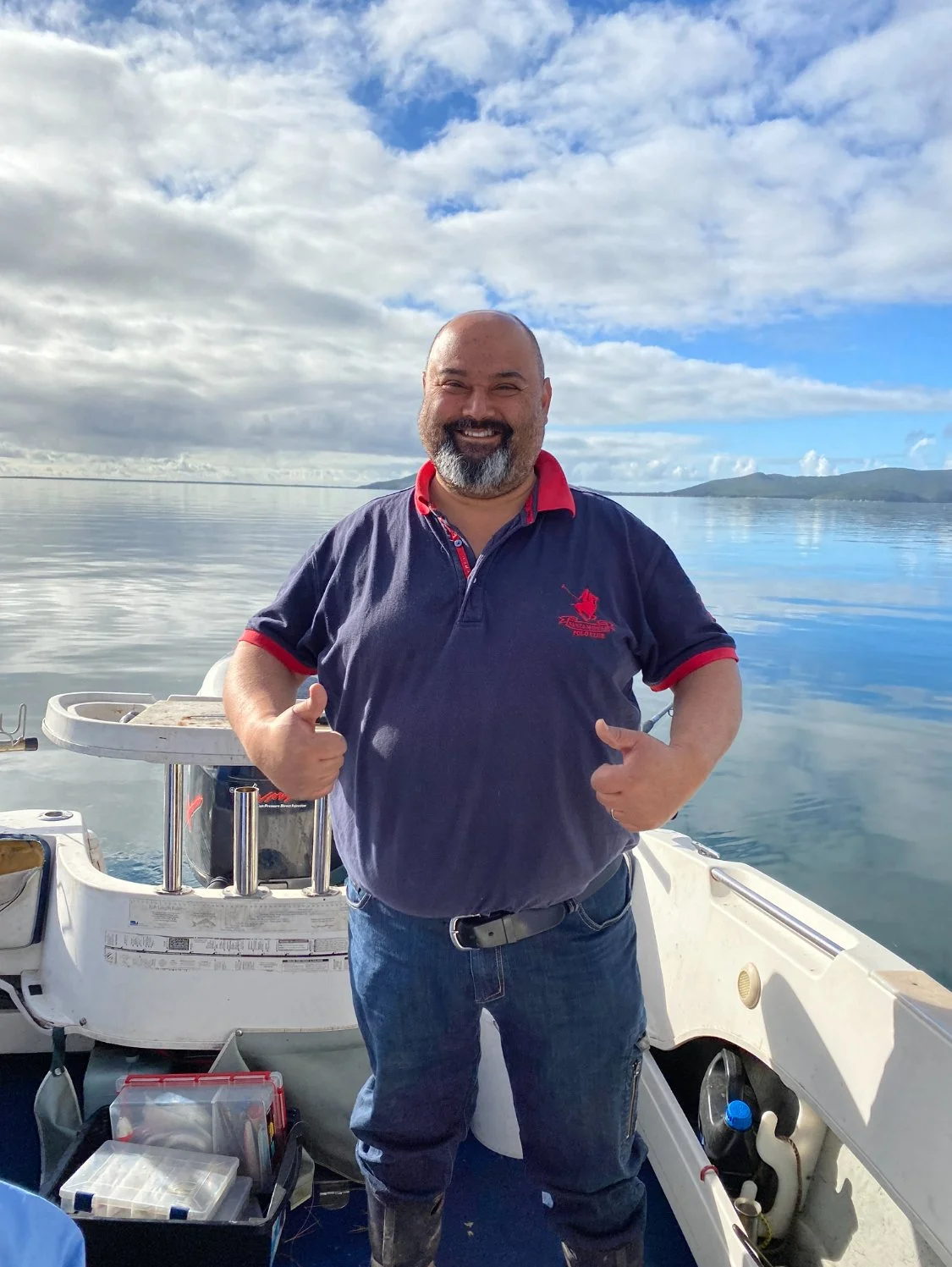 A man with a beard, smiling and giving a thumbs-up while standing on a boat in a calm body of water. The sky is partly cloudy with some blue patches, and there are mountains or hills in the background.