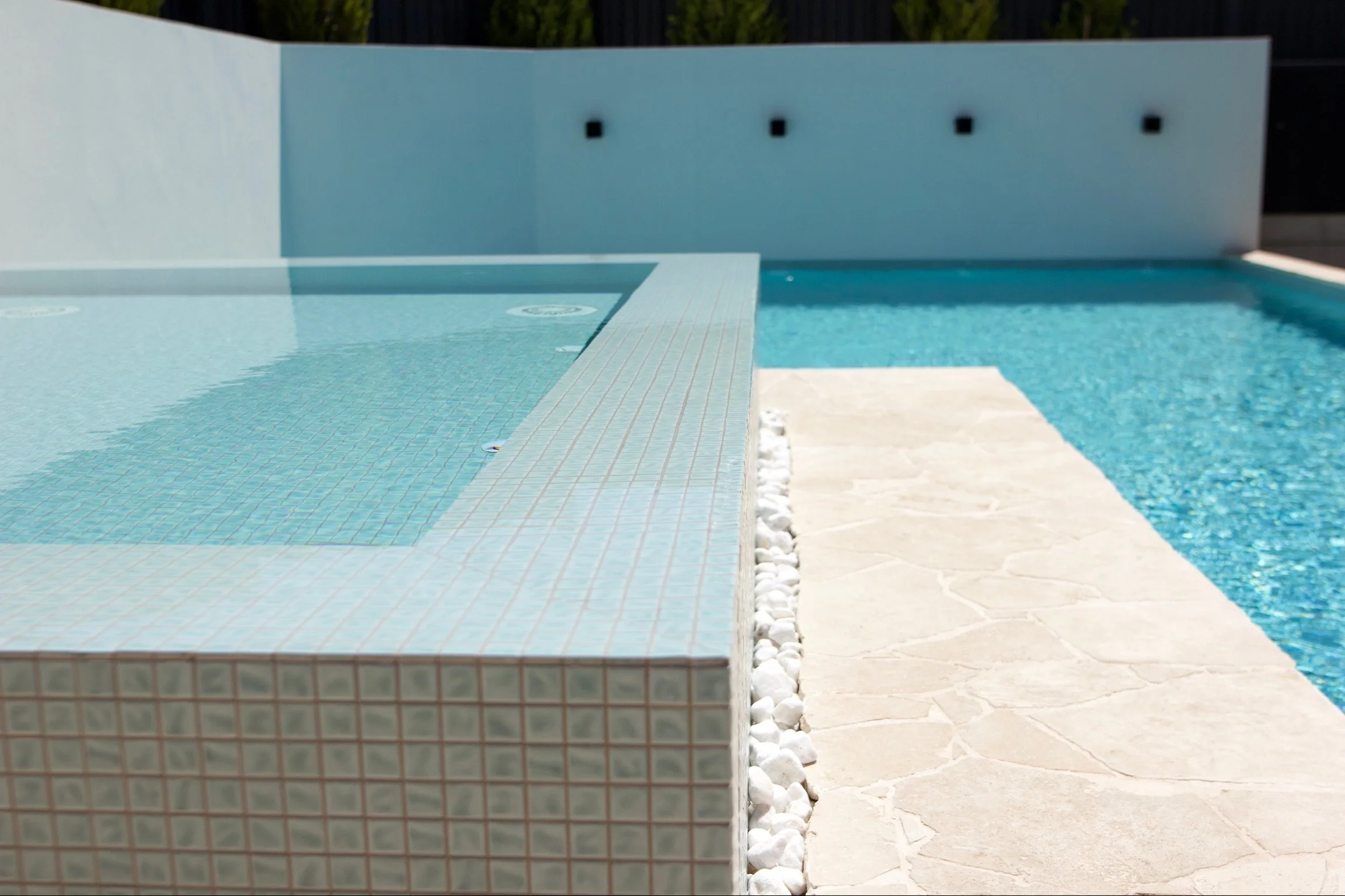 Close-up of a modern swimming pool with light blue tiles, a white stone deck, and a white wall with black water jets in the background.
