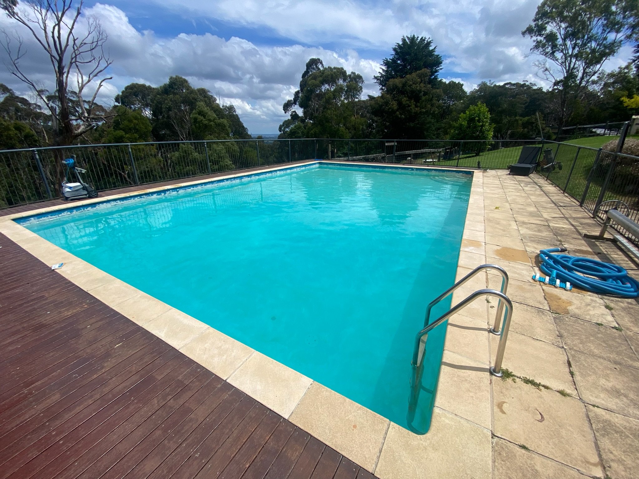 An outdoor swimming pool surrounded by a stone and wooden deck, with a grassy area and trees in the background. The pool has metal railings at the steps, a blue pool hose coiled on the ground, and a black lounge chair. The sky is partly cloudy with blue patches.