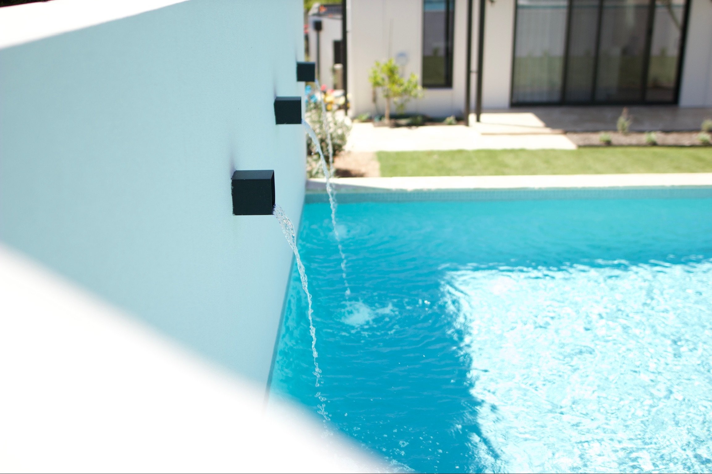 Close-up of a modern outdoor pool feature with water flowing from three small black spouts onto the pool's water surface, with a house and garden in the background.