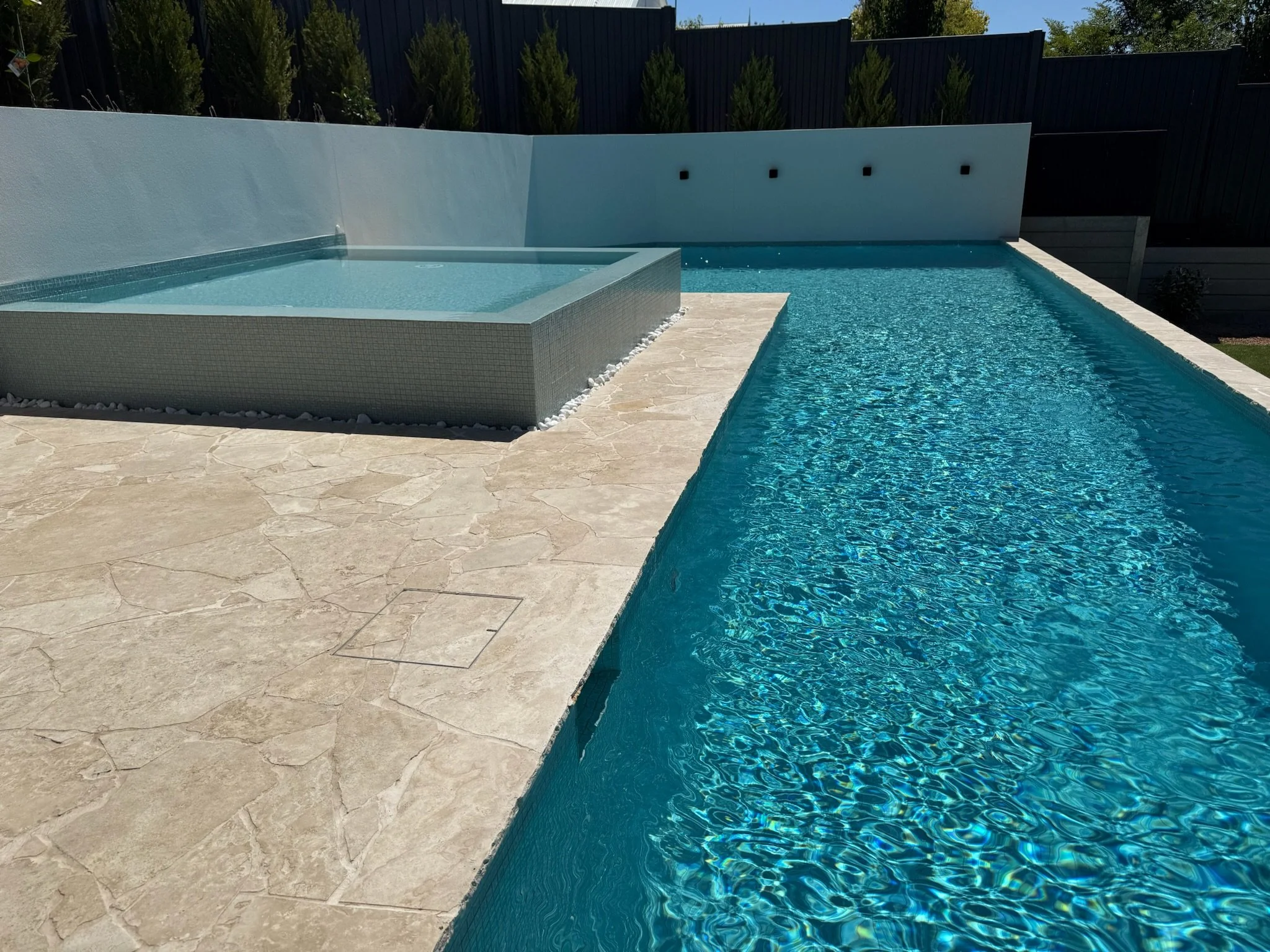 A modern swimming pool with a raised jacuzzi on a beige stone deck, surrounded by a white wall and tall evergreen trees in the background.