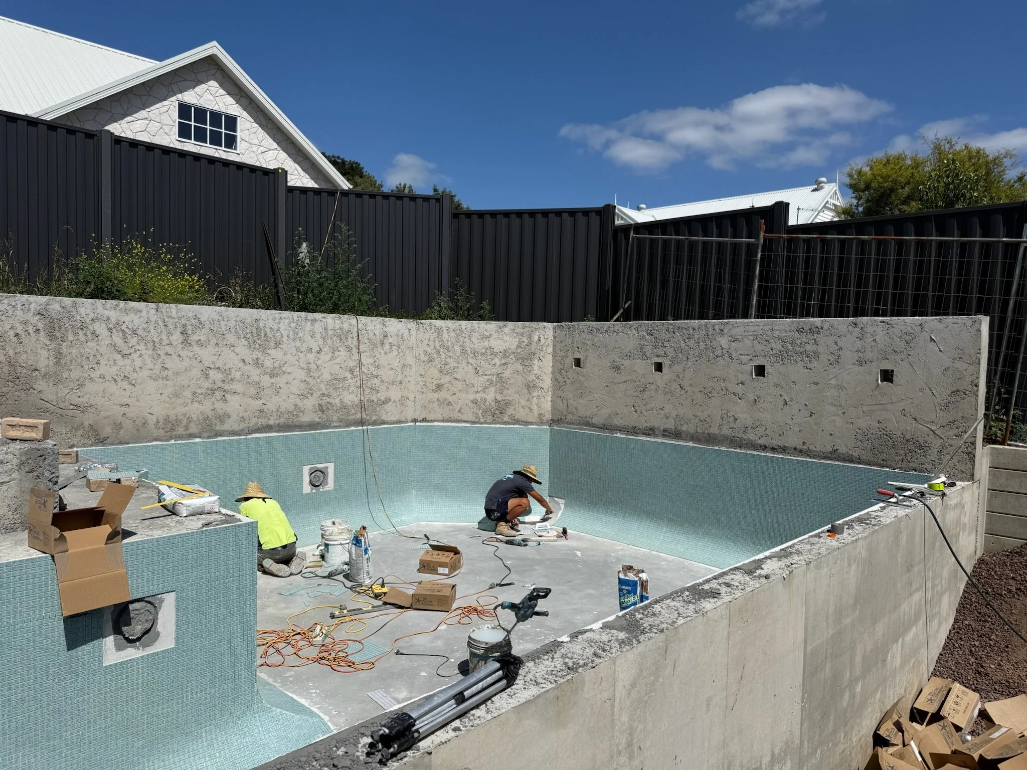 Construction workers installing tiles in a swimming pool outdoors under a blue sky with some clouds. The pool has light blue tiles and is surrounded by a concrete wall with small square openings. There are construction tools, boxes, and cords on the ground.