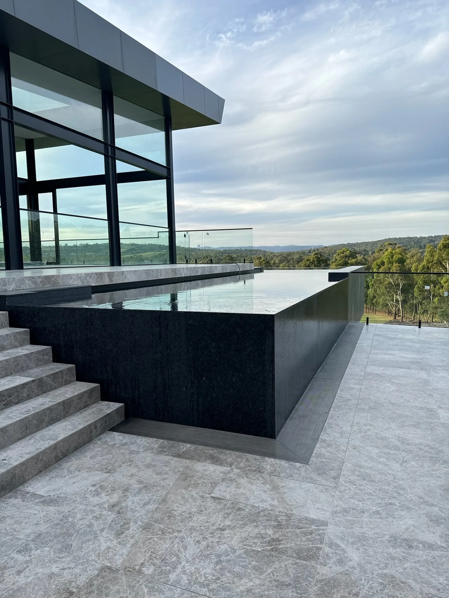 Modern building with large glass windows and an infinity pool overlooking green trees and a cloudy sky.