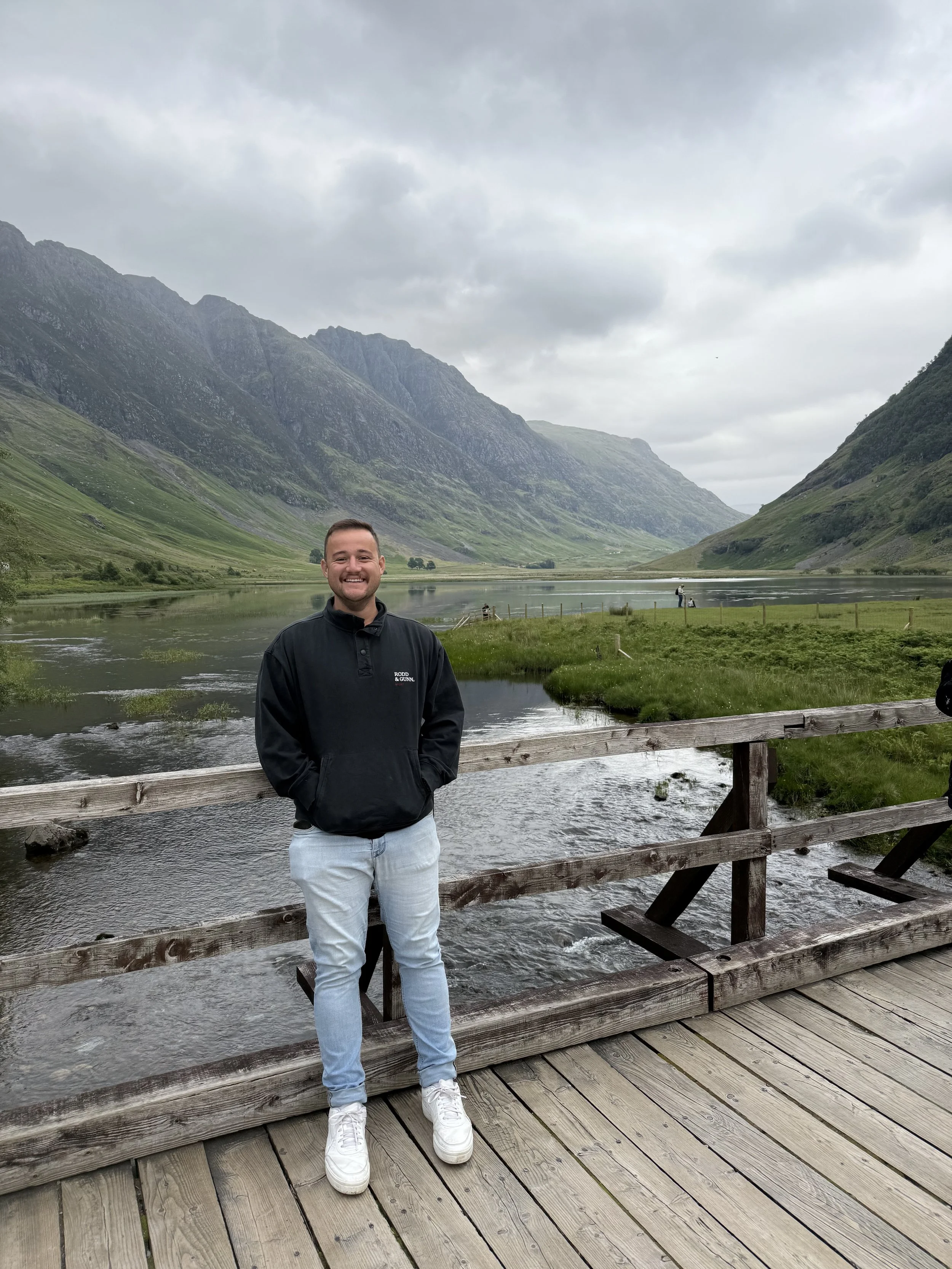 A man standing on a wooden bridge near a river with mountains and cloudy sky in the background.
