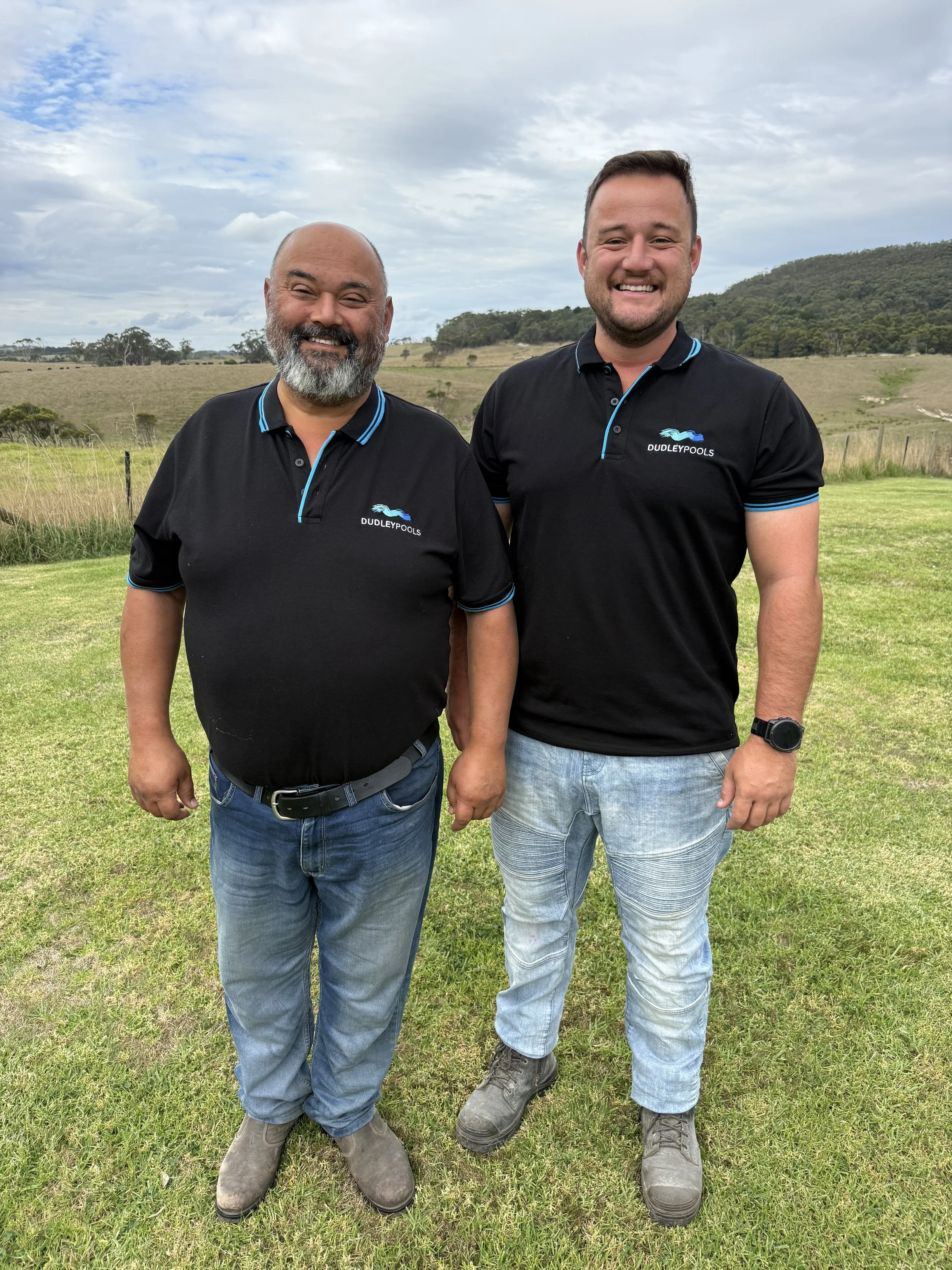 Two men standing outdoors on grass, smiling, wearing black polo shirts with the Dubleypools logo, with a rural landscape and cloudy sky in the background.