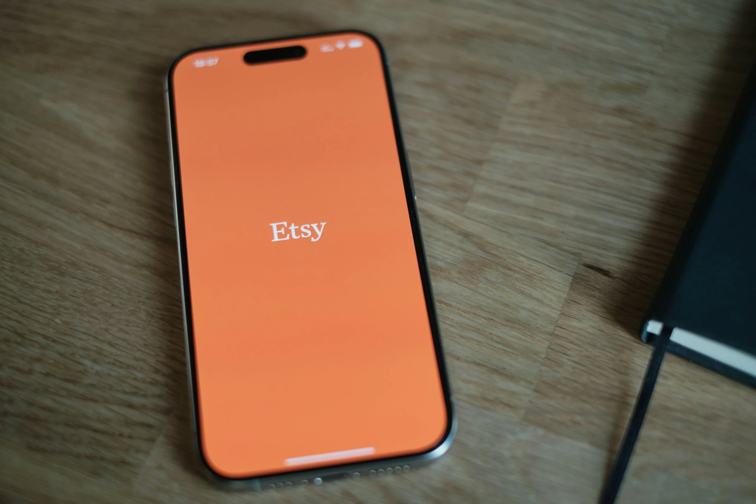 An iPhone with an orange screen displaying the Etsy logo lies on a wooden surface.