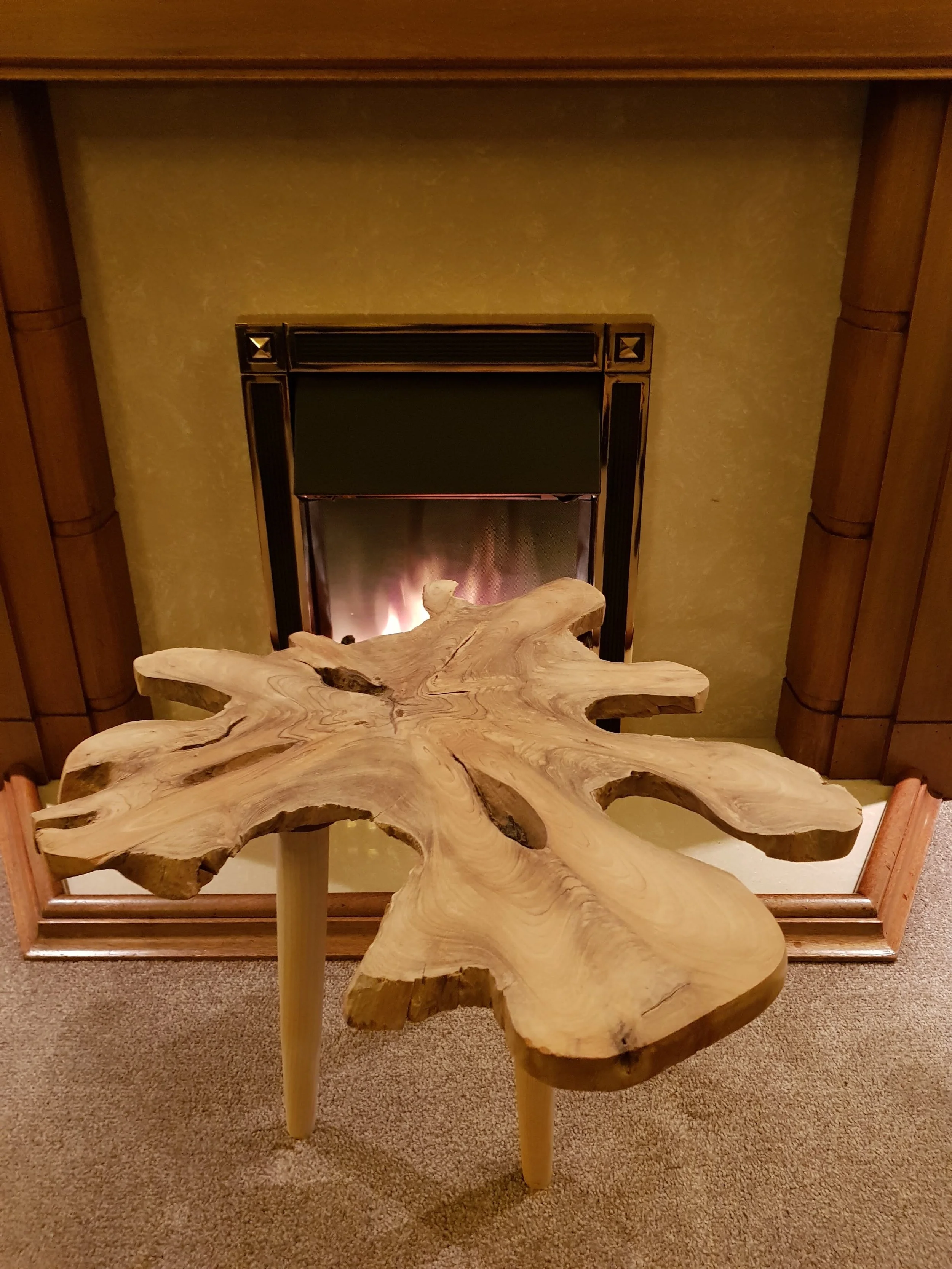 A unique wooden table with a natural, uneven shape and exposed wood grains sits in front of a fireplace with a yellow wall background.