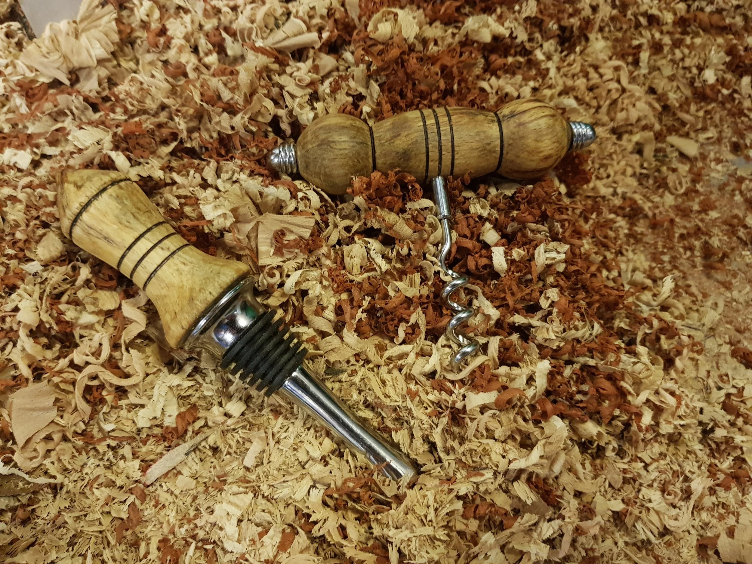 A corkscrew and bottle stop with a wooden handle lying on a bed of wood shavings.