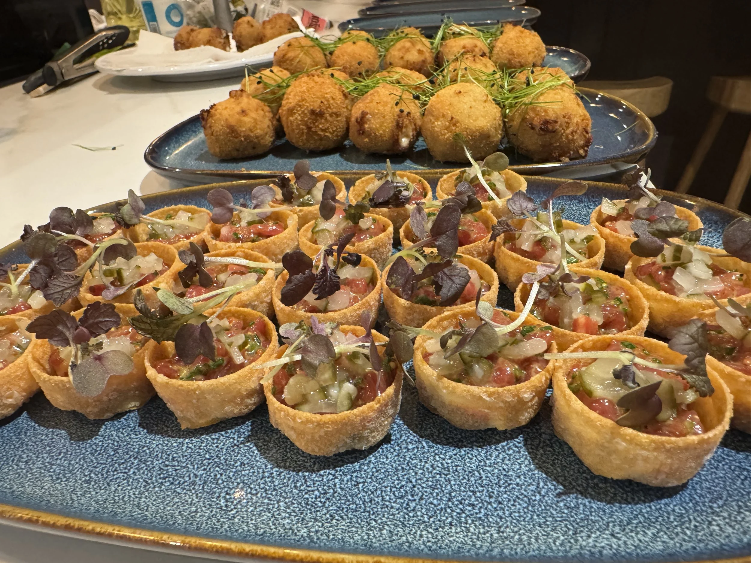 Tray of bite-sized appetizer scoops topped with microgreens and a second tray of breaded, fried balls garnished with microgreens in the background.