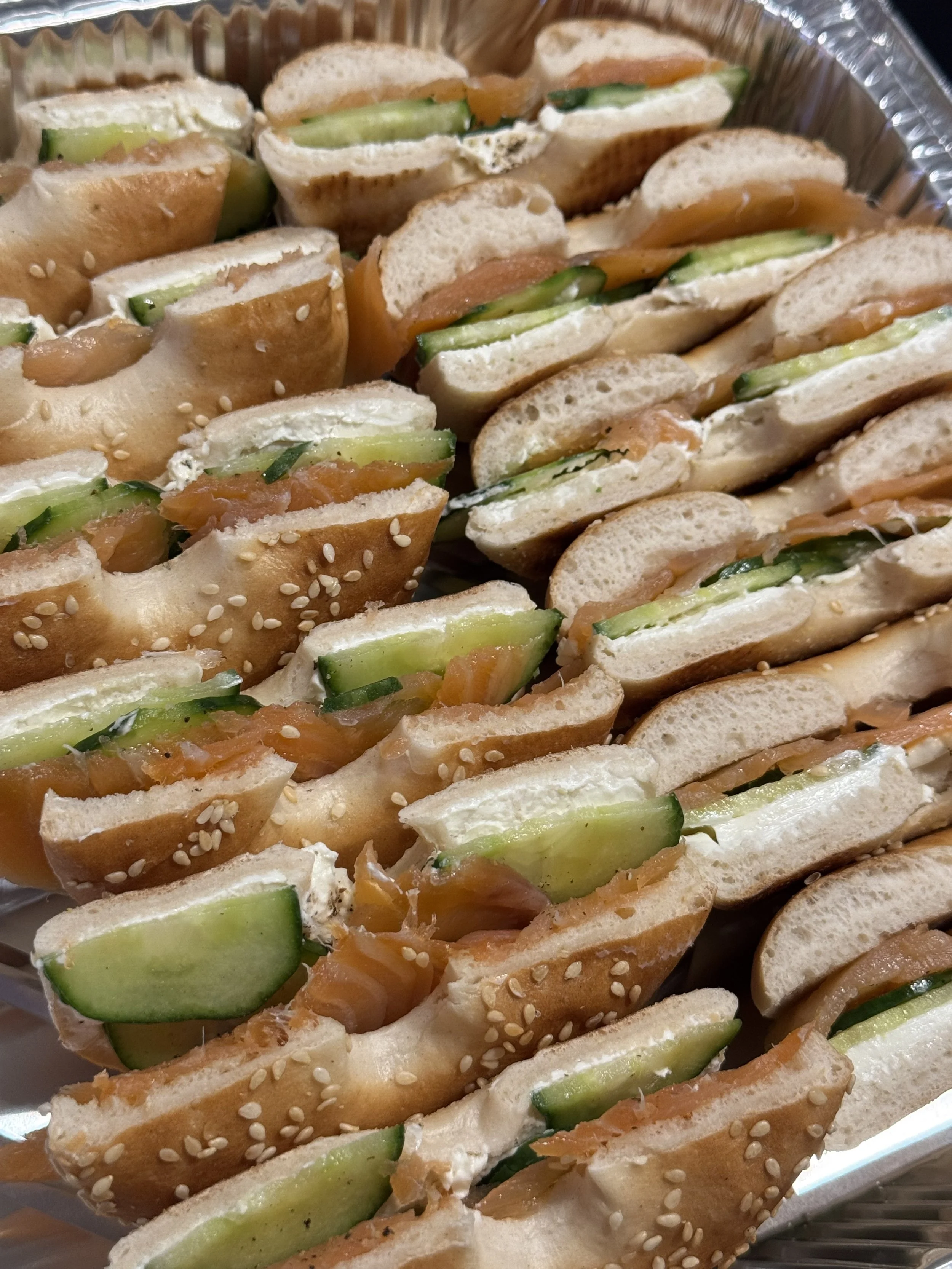 Assorted mini sandwiches with smoked salmon, cucumber, and cream cheese on sesame seed baguette slices, arranged in a tray.